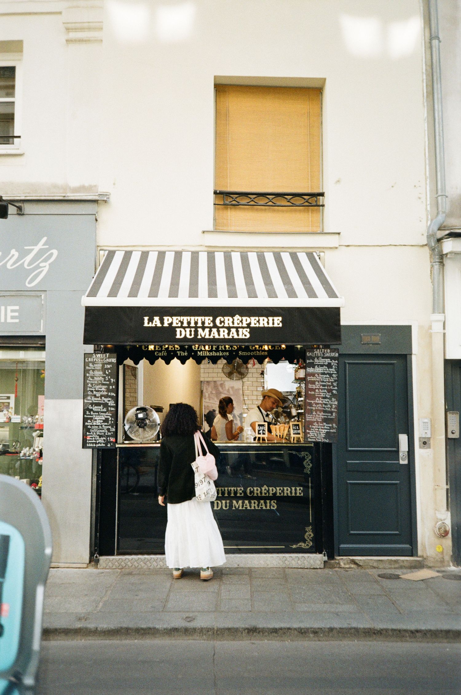 Woman in a long white skirt stands at the counter of La Petite Crêperie du Marais, a small Paris crepe shop with a striped awning.