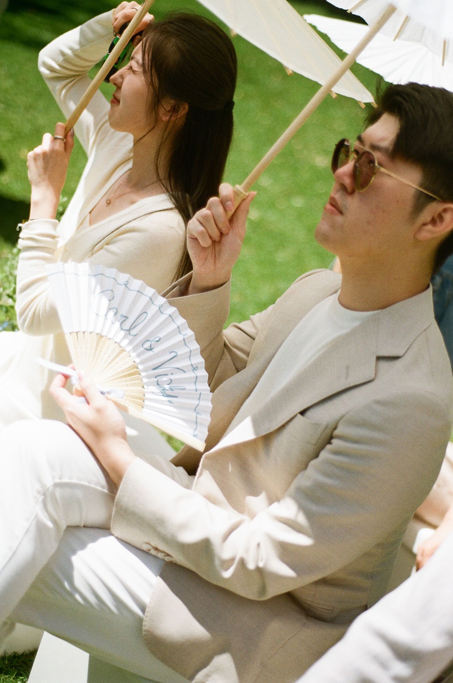 Man in sunglasses and beige suit holds a paper parasol beside a woman with a white fan at an outdoor garden wedding ceremony.