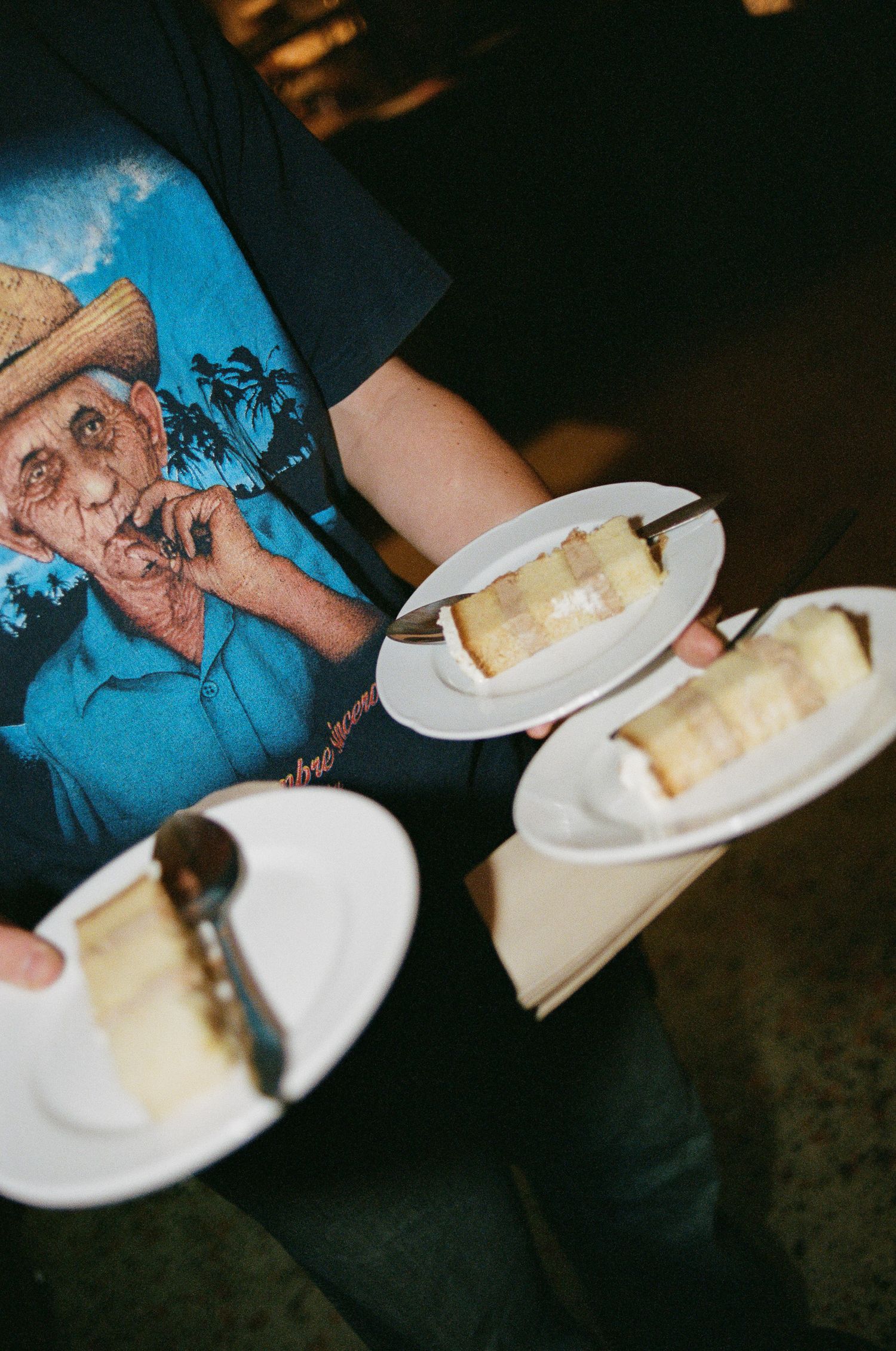 Person in a graphic tee holding three white plates with slices of layered cake and forks, serving dessert at a wedding or party.