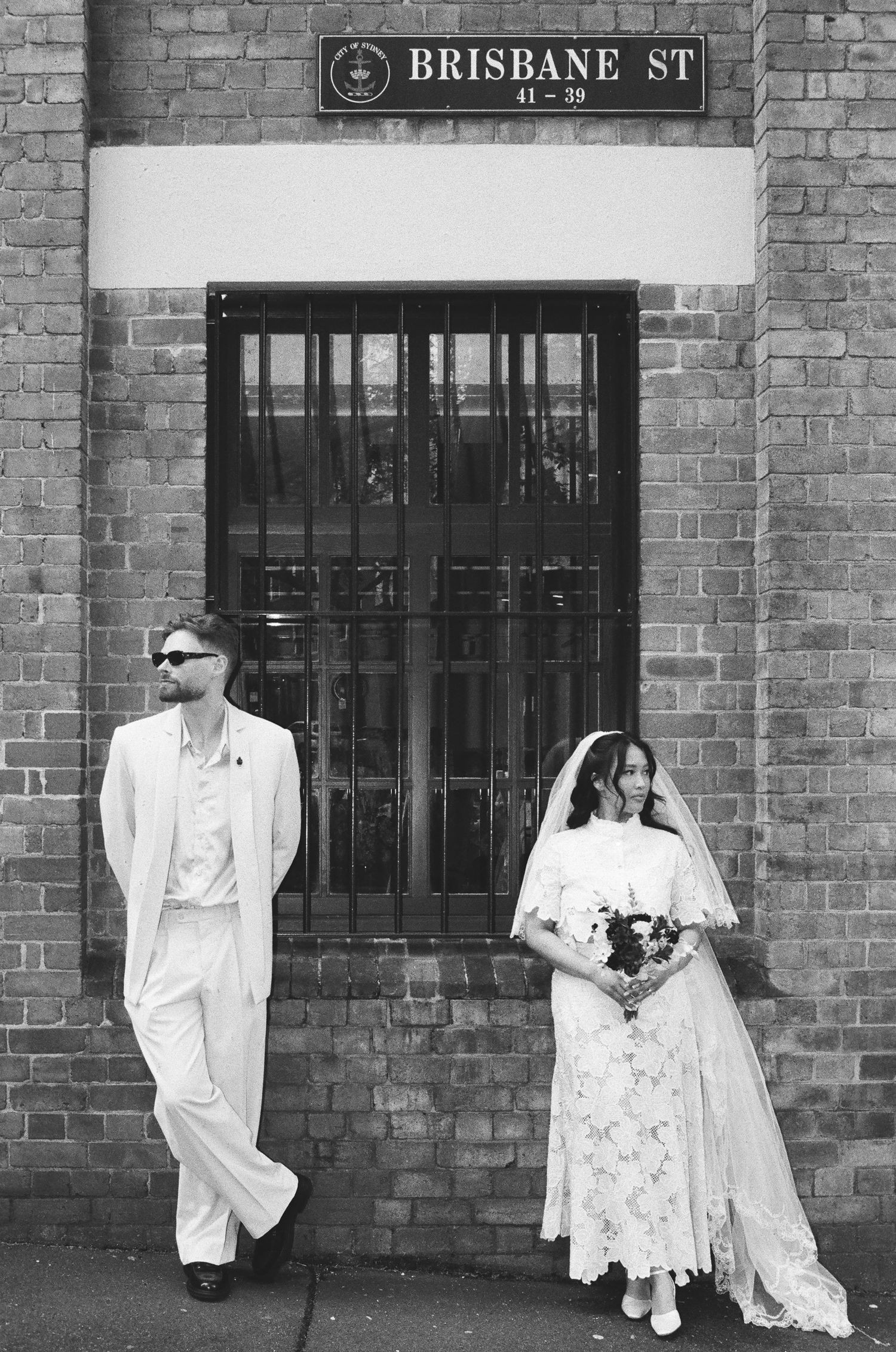 Black and white wedding portrait of a groom in a light suit and sunglasses and a bride in a lace dress and veil by a brick wall.
