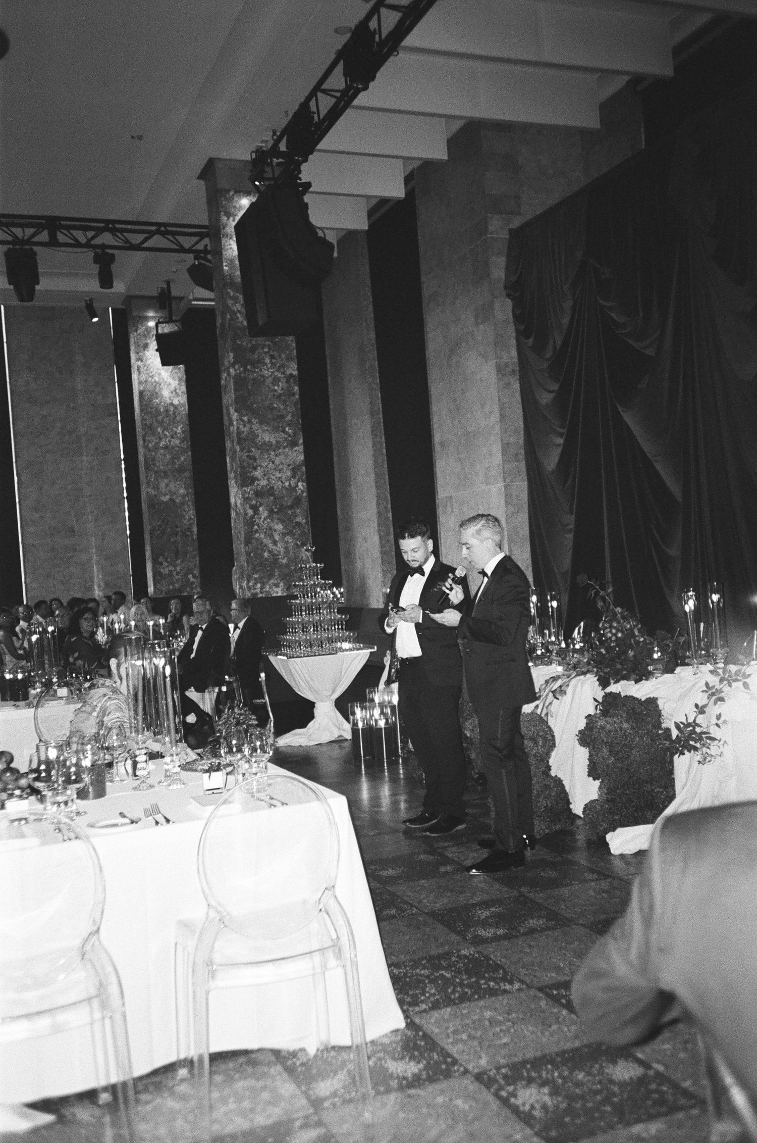 Two men in formal attire stand speaking in an elegant wedding reception hall with marble columns, draped tables and a champagne tower.