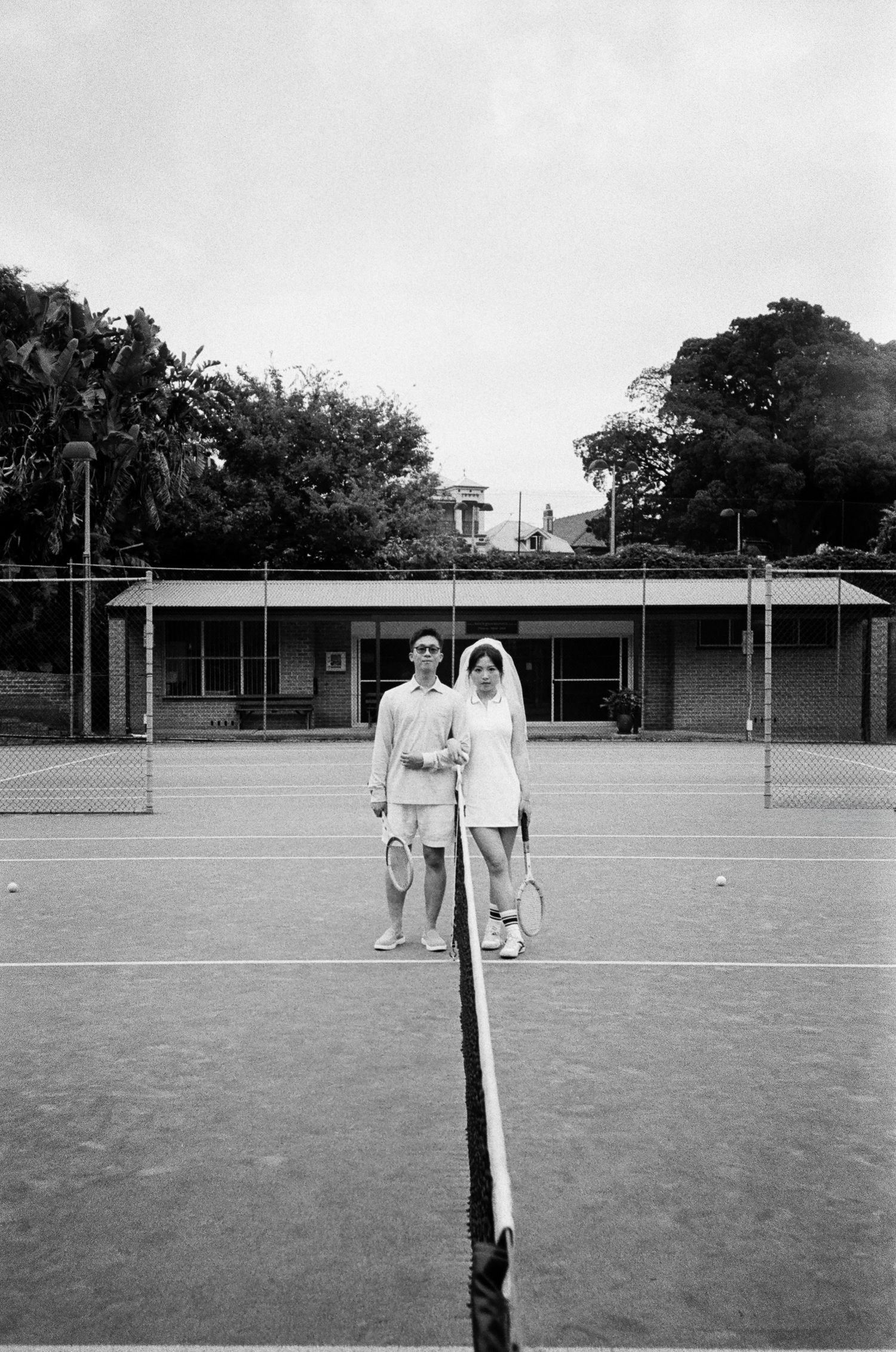 Black and white photo of a couple in tennis whites holding rackets on a court, the woman wearing a bridal veil, standing behind the net.