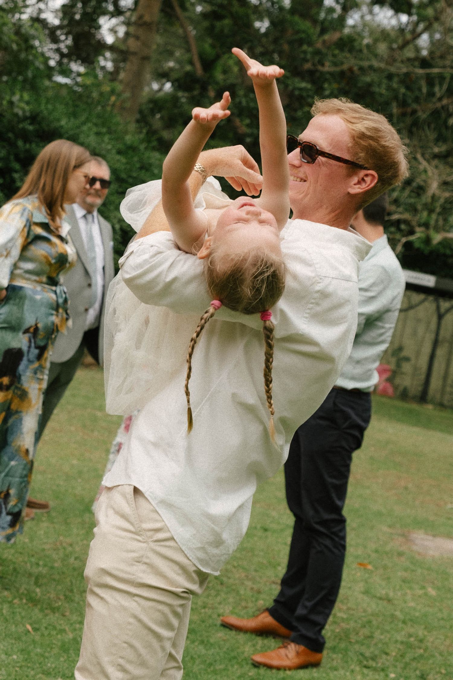 A person in white clothing drinks from a glass bottle while standing outdoors on grass during a social gathering.