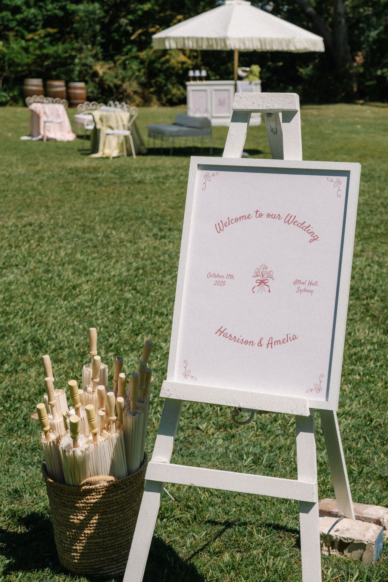 White wedding welcome sign on an easel with parasol umbrellas and chairs set up on a sunny lawn for an outdoor ceremony.