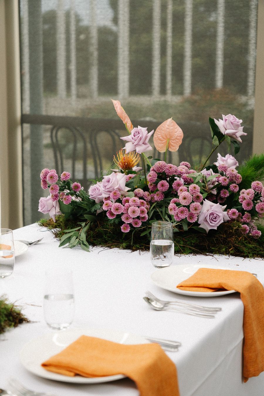An elegant table setting with pink floral centerpiece and orange napkins on a white tablecloth by a window.