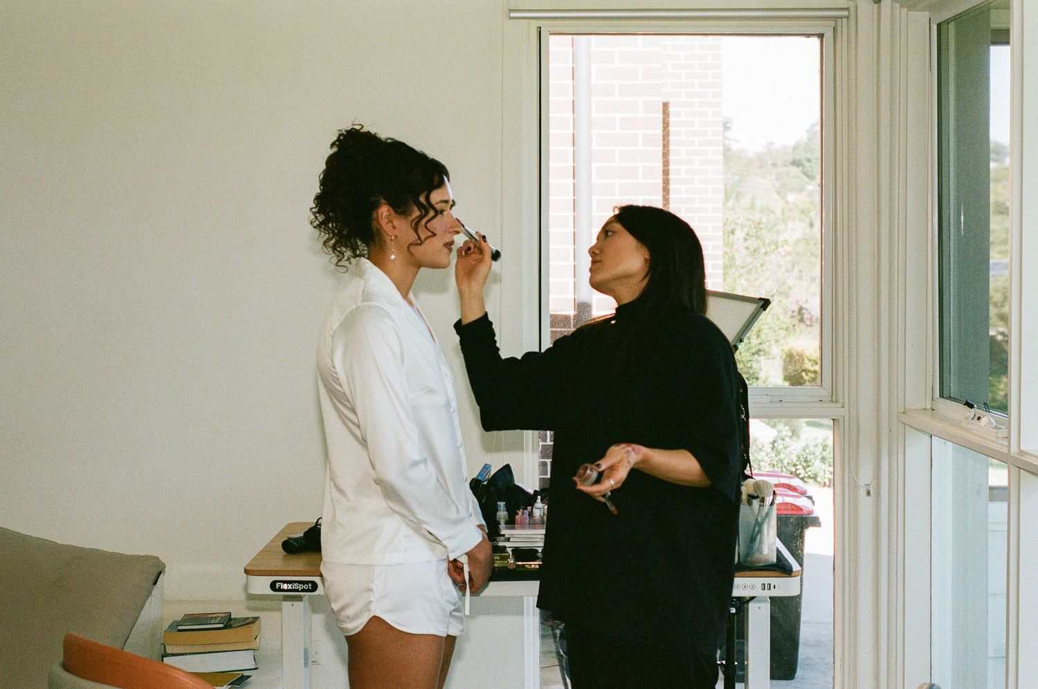 A makeup artist in black applies lipstick to a woman in a white robe beside a bright window in a sunlit room.
