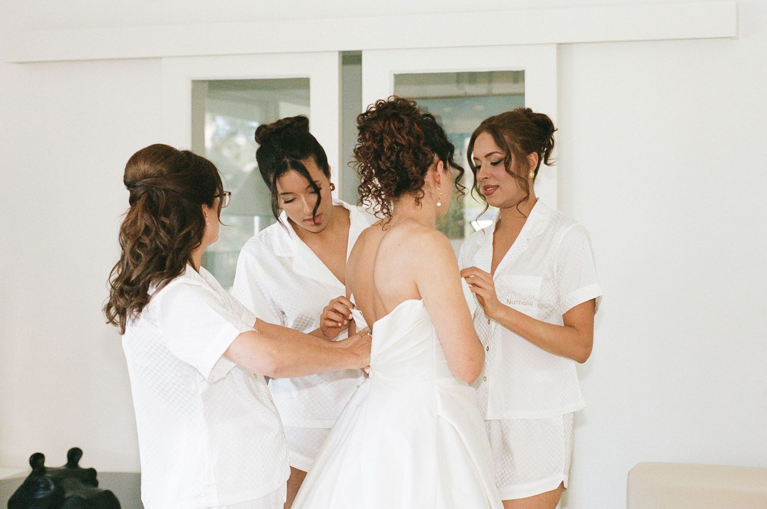 Bride in a white wedding dress gets help from three bridesmaids in white robes while getting ready in a bright room.