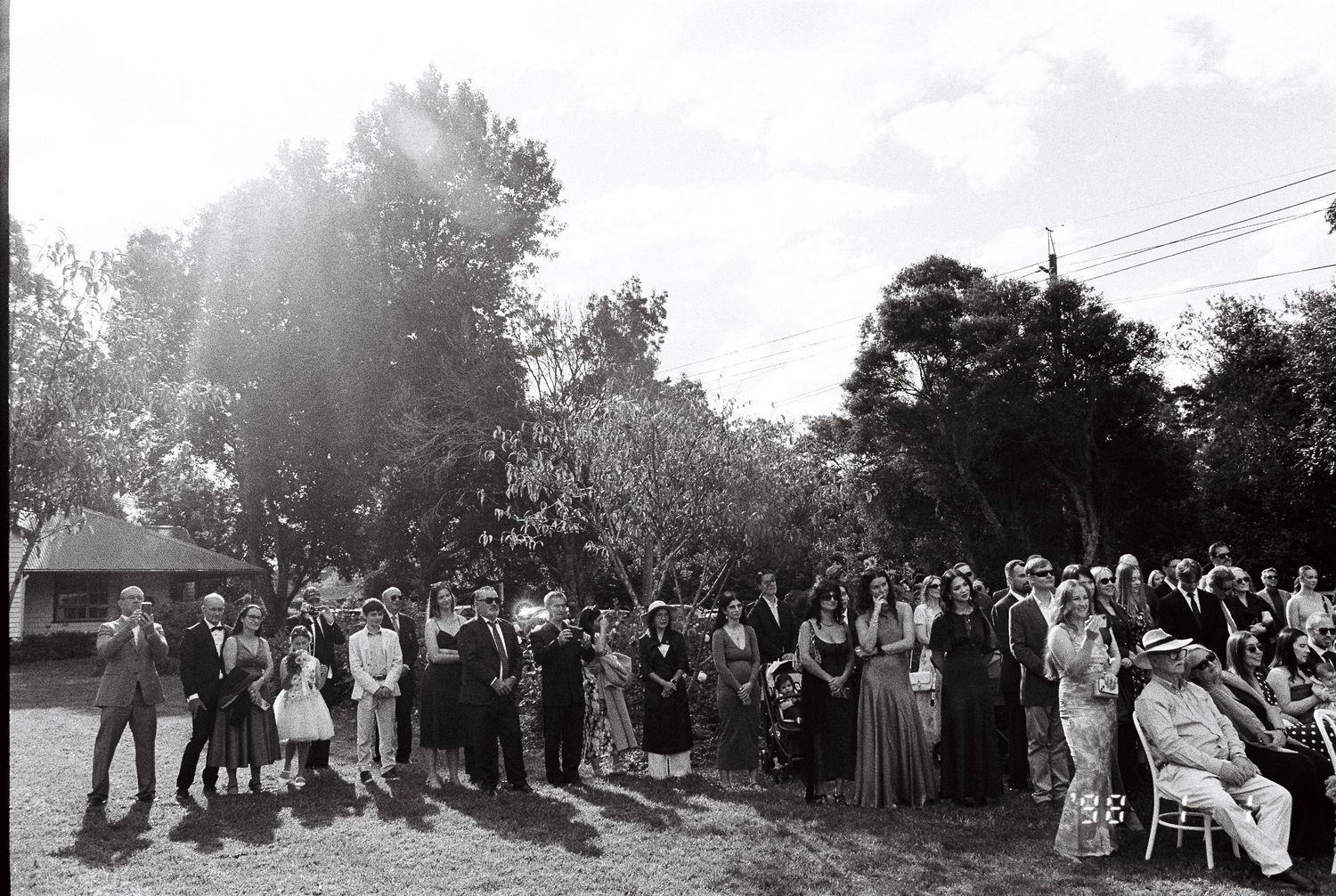 Black and white photo of wedding guests standing and seated on a grassy lawn beneath tall trees during an outdoor ceremony.