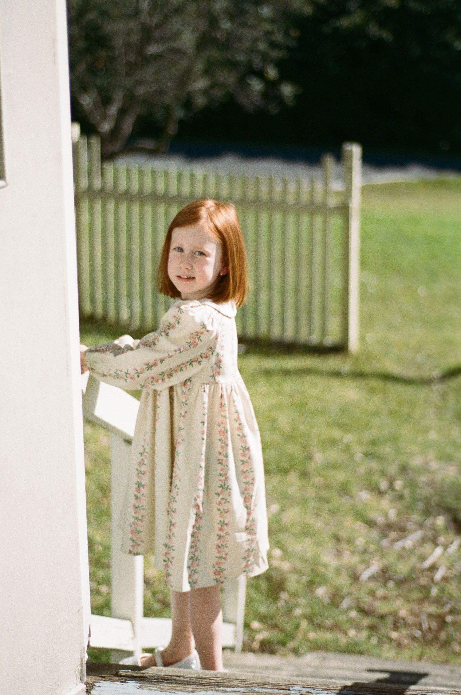 A young red-haired girl in a floral dress stands barefoot by a white fence, smiling outdoors on a sunny day.