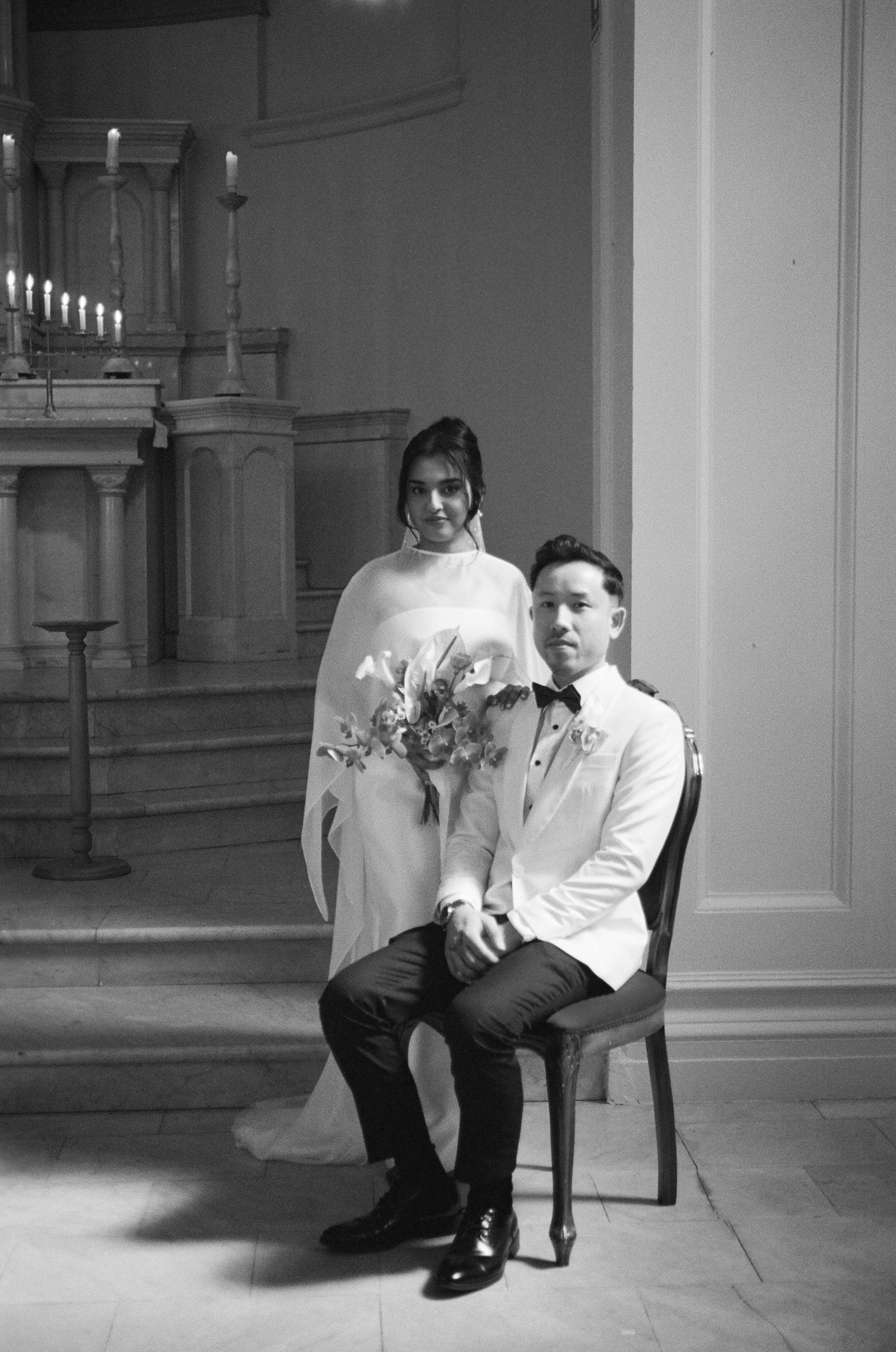 Black and white wedding portrait of a bride in a flowing veil holding a bouquet standing behind a seated groom in a white tuxedo jacket.