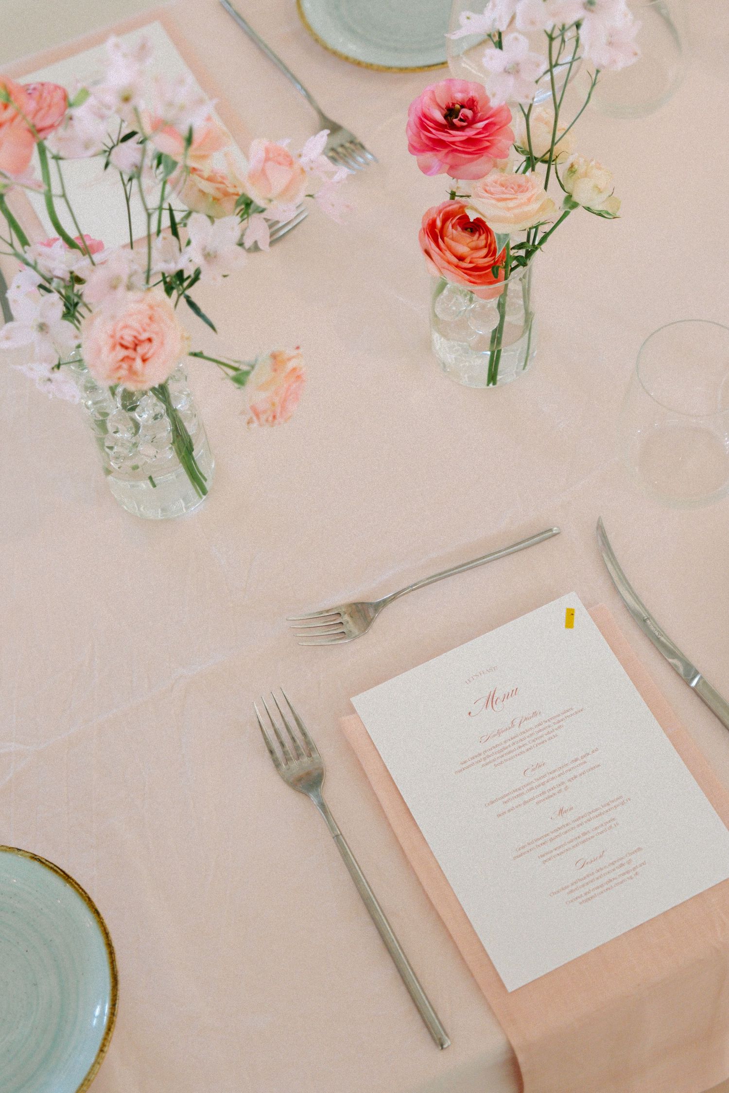 Elegant table setting with pink roses in glass vases, menu card, and place settings on a pale pink tablecloth.