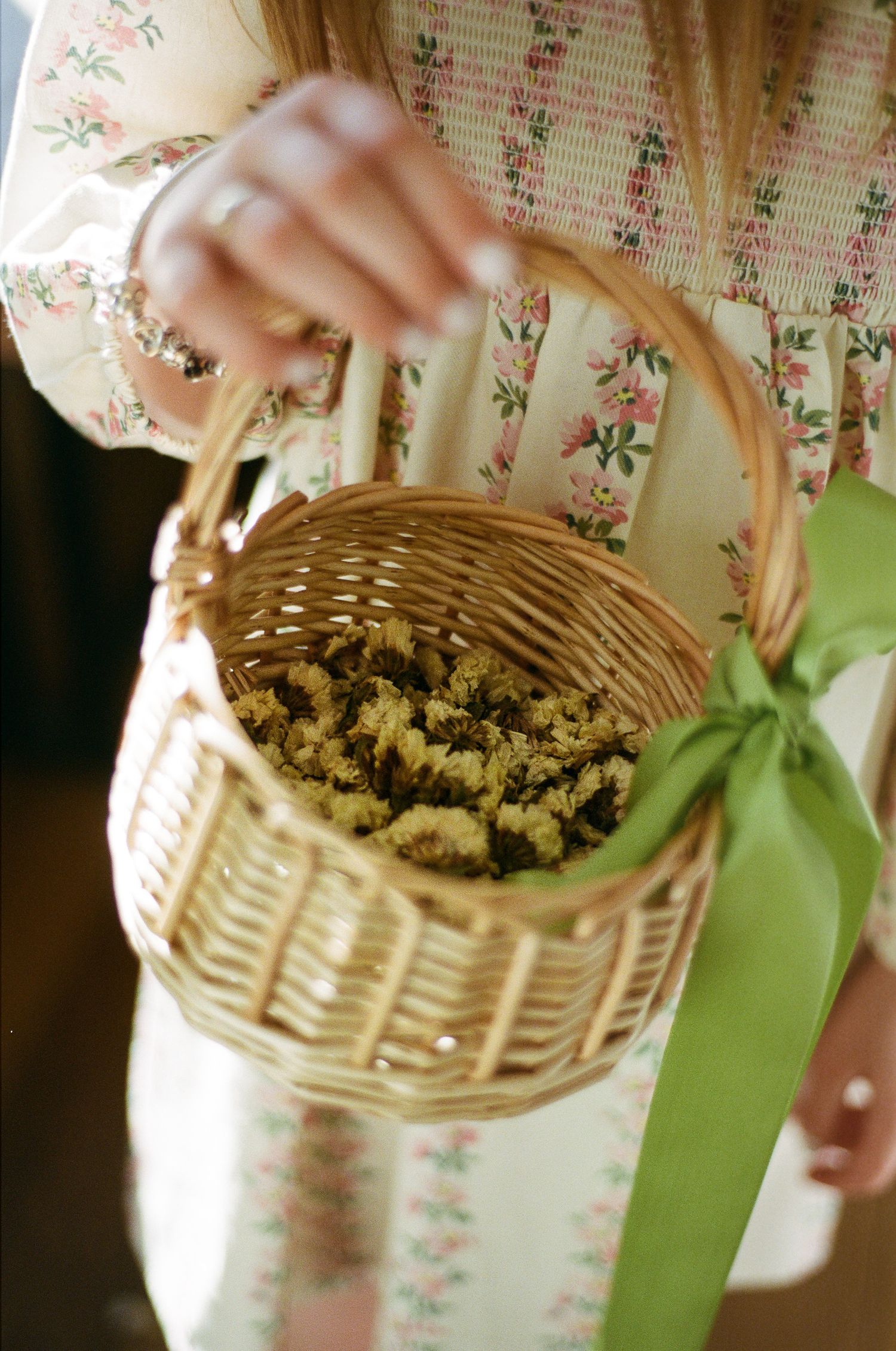 Woman in a floral smocked dress holding a wicker basket of dried flowers tied with a green ribbon.