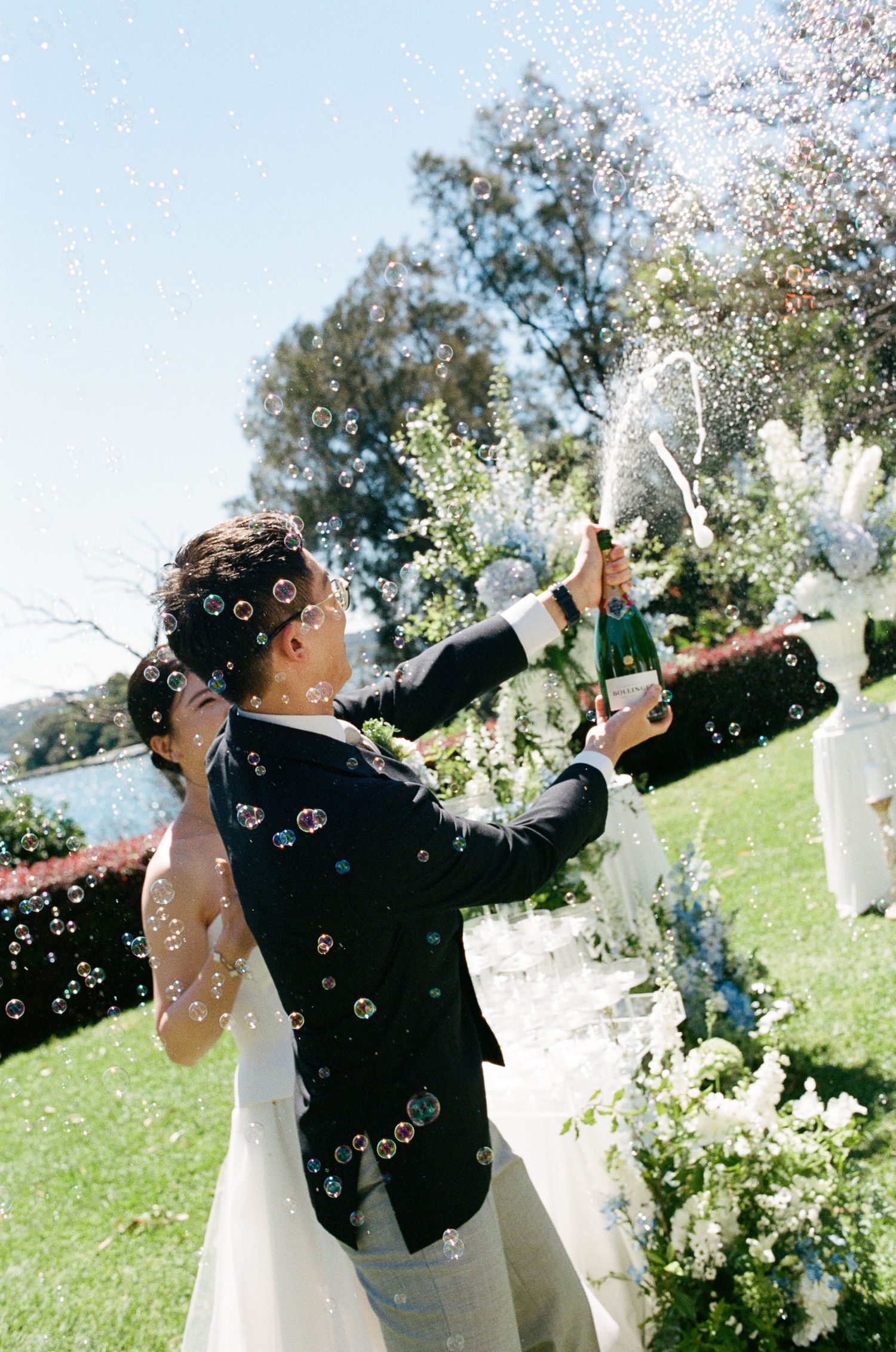 Groom in a dark suit sprays champagne beside a bride in a white gown as bubbles float through the air at an outdoor garden wedding.
