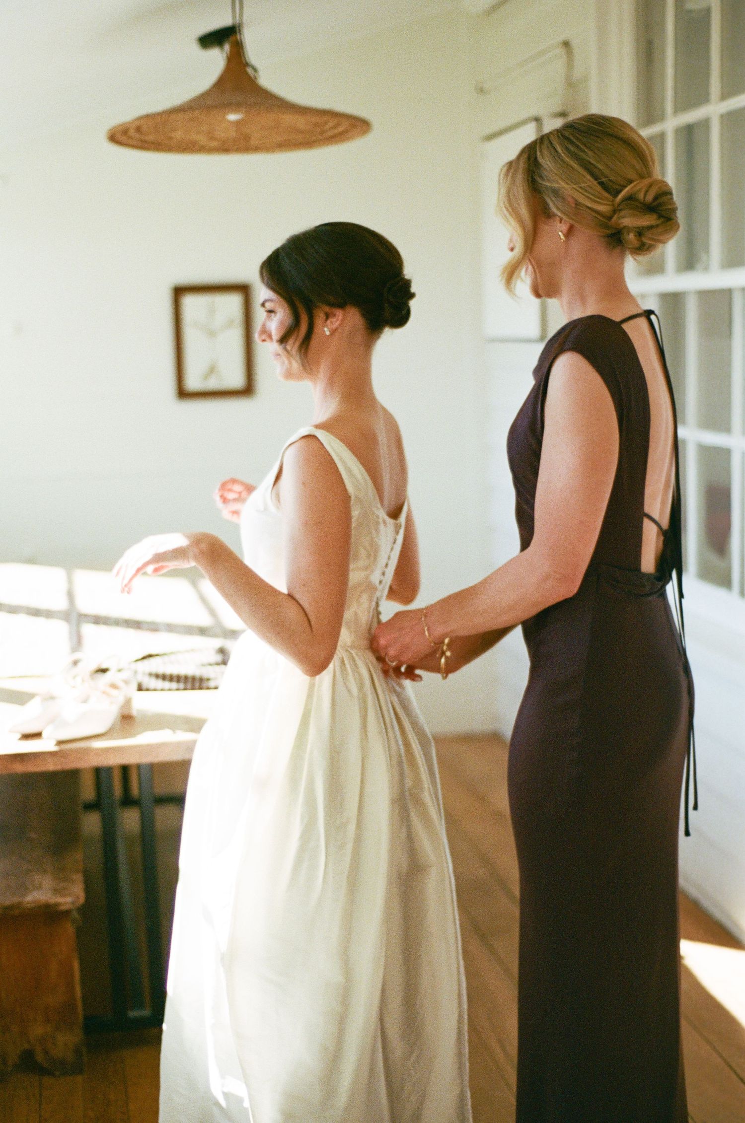 Bride in elegant white gown being helped by bridesmaid in black dress during wedding preparations.