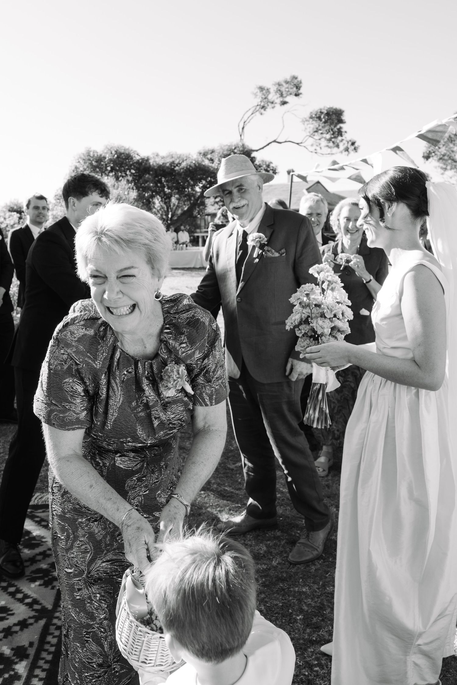 Black and white photo of wedding guests smiling as a child reaches into a basket at an outdoor ceremony.