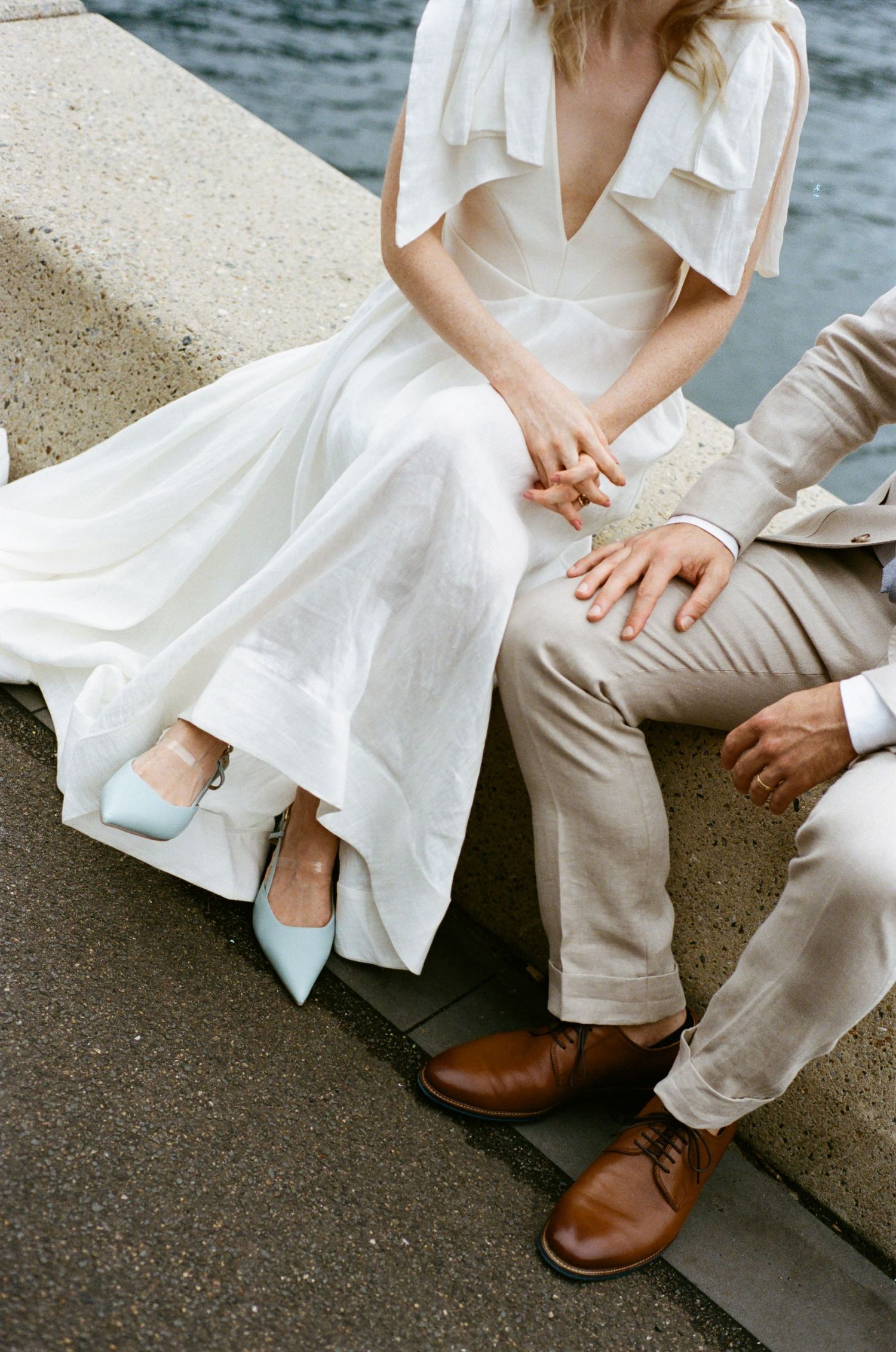 Close-up of two people sitting together wearing white and beige clothing with brown leather shoes by the water.