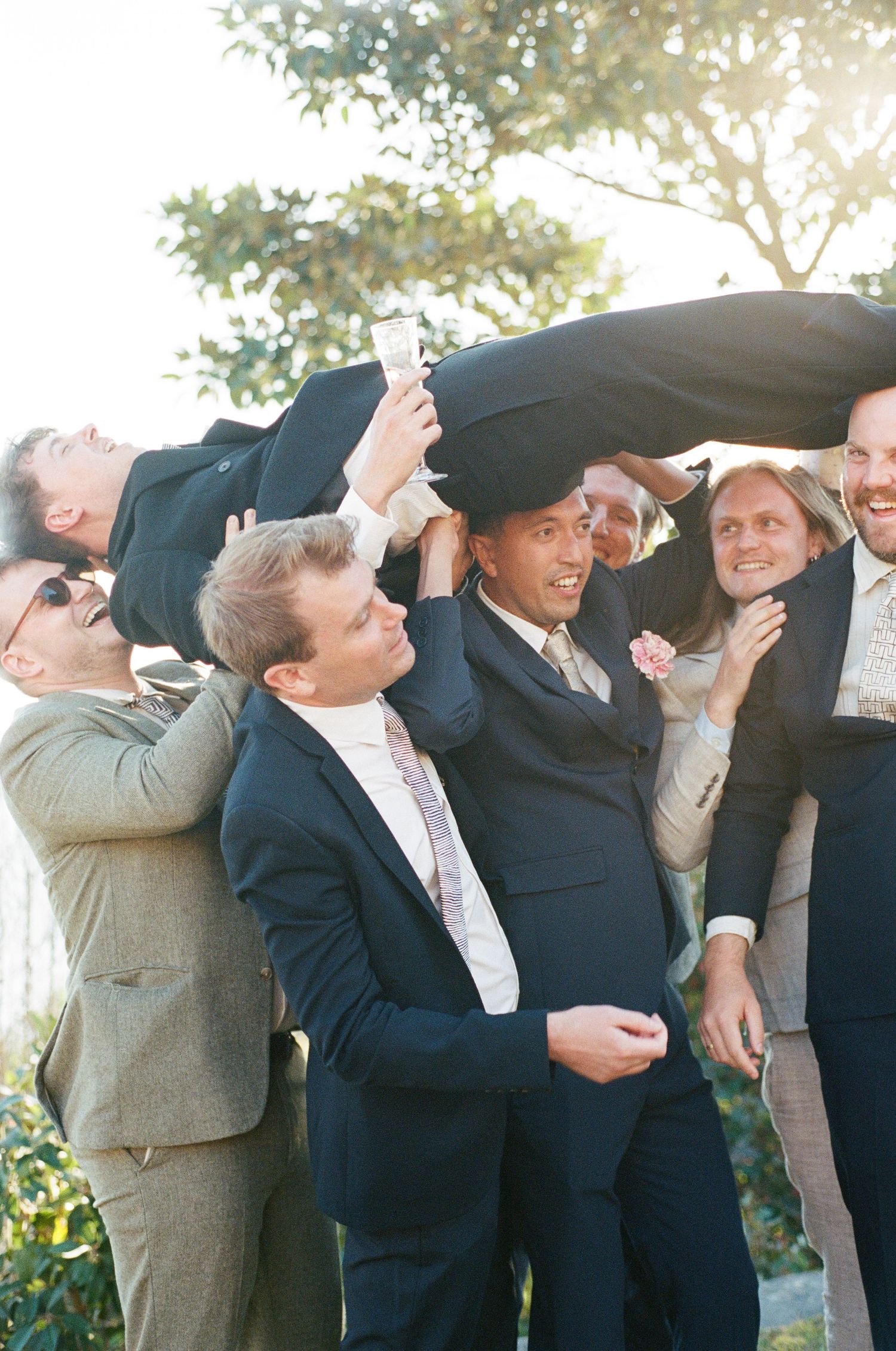 Groomsmen lifting and celebrating groom in navy suit at outdoor wedding reception in sunlight.