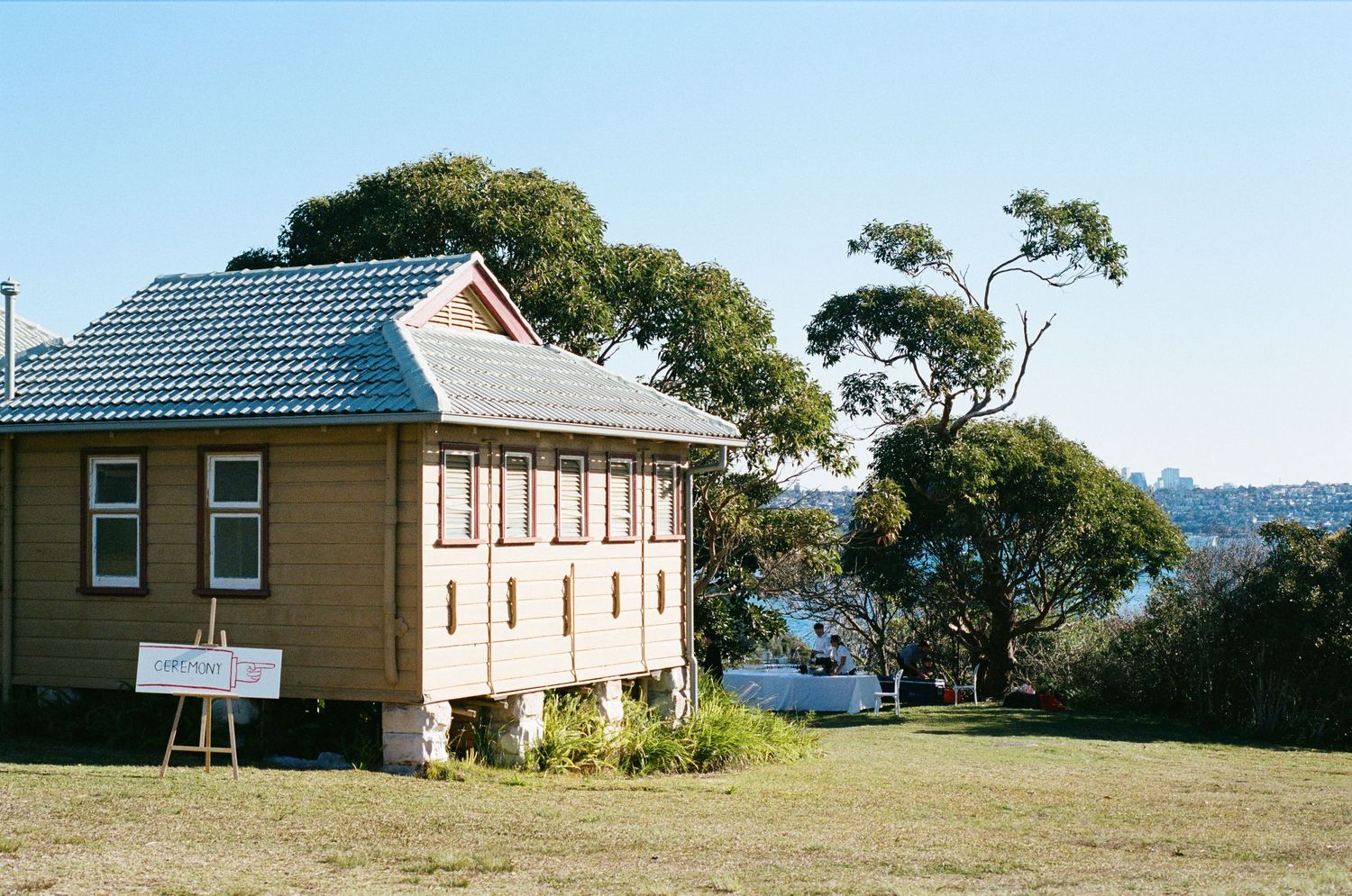 Historic yellow wooden building on stilts with blue roof surrounded by eucalyptus trees and green lawn near Sydney Harbour.