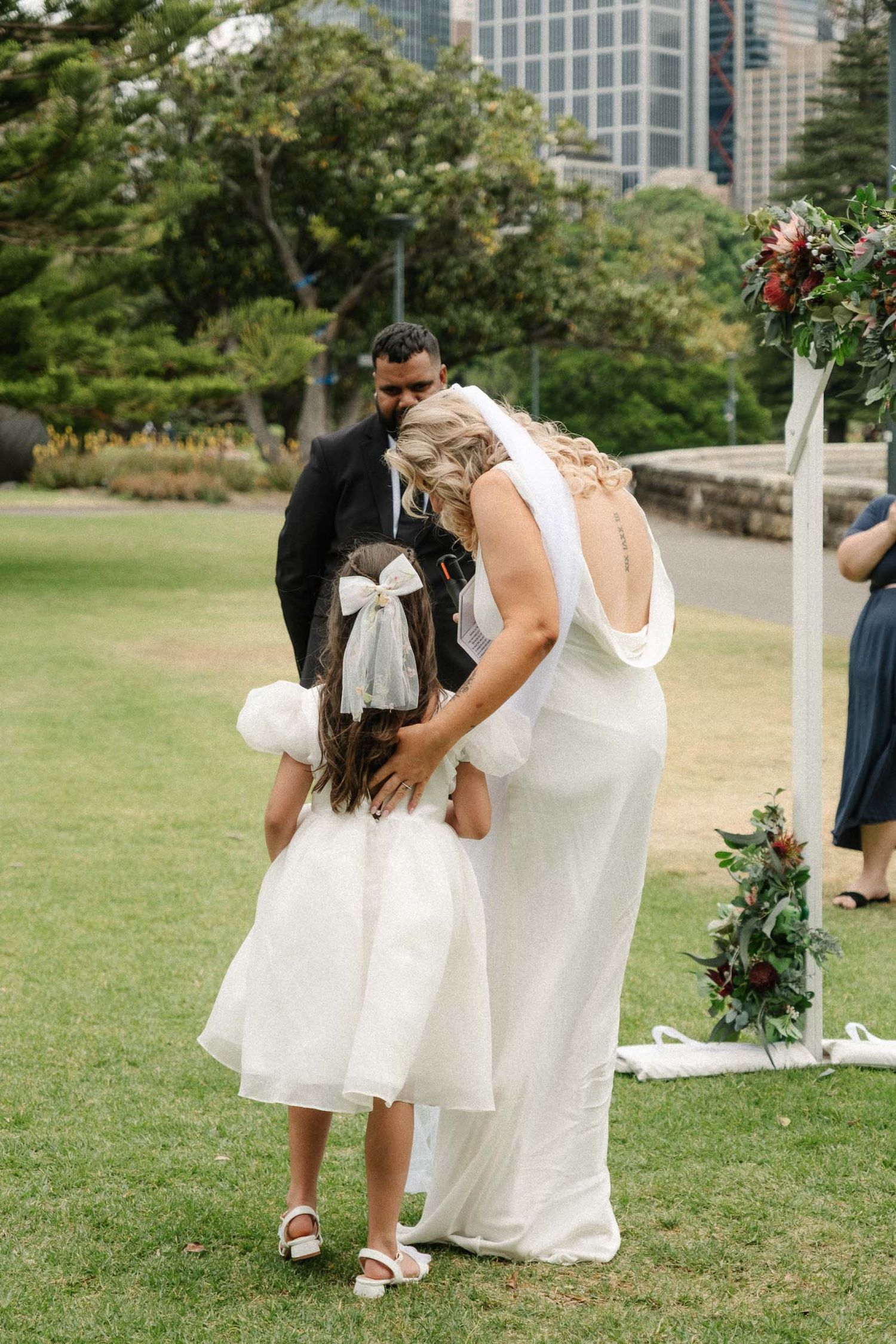 A wedding couple embraces on a grassy lawn with city buildings visible in the background on a sunny day.