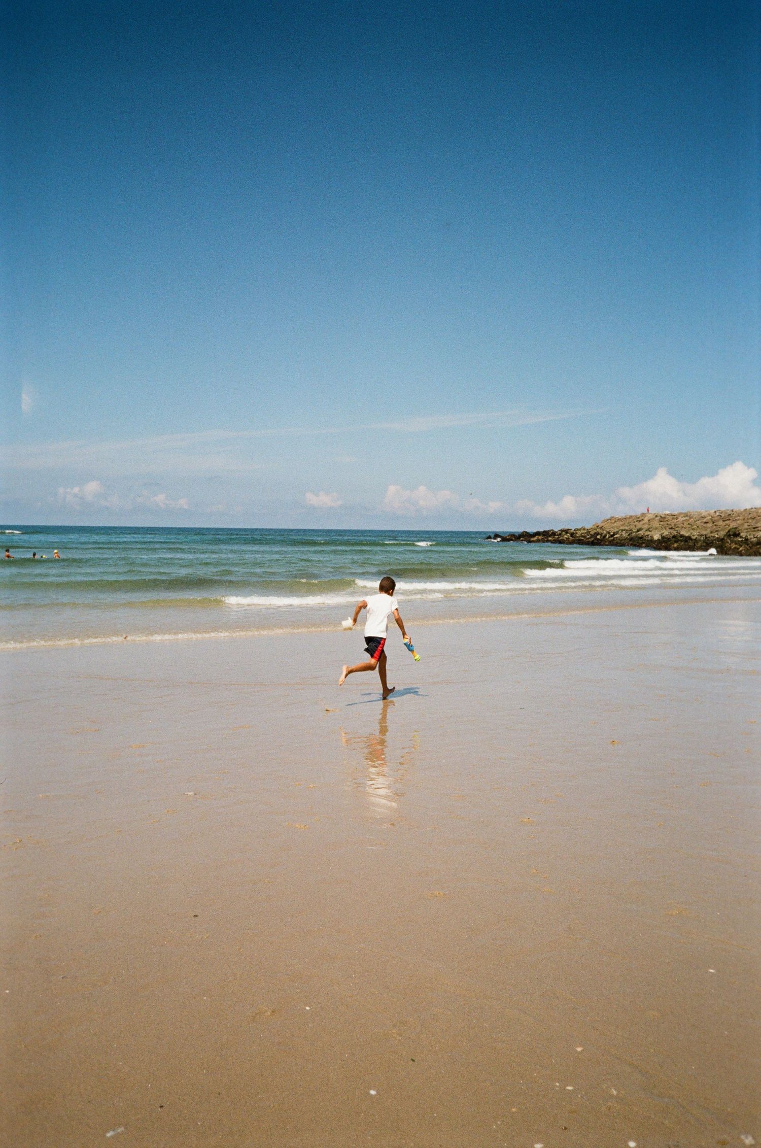 A person in a white shirt runs across wet sand toward the waves on a sunny beach with blue sky and rocky coastline.