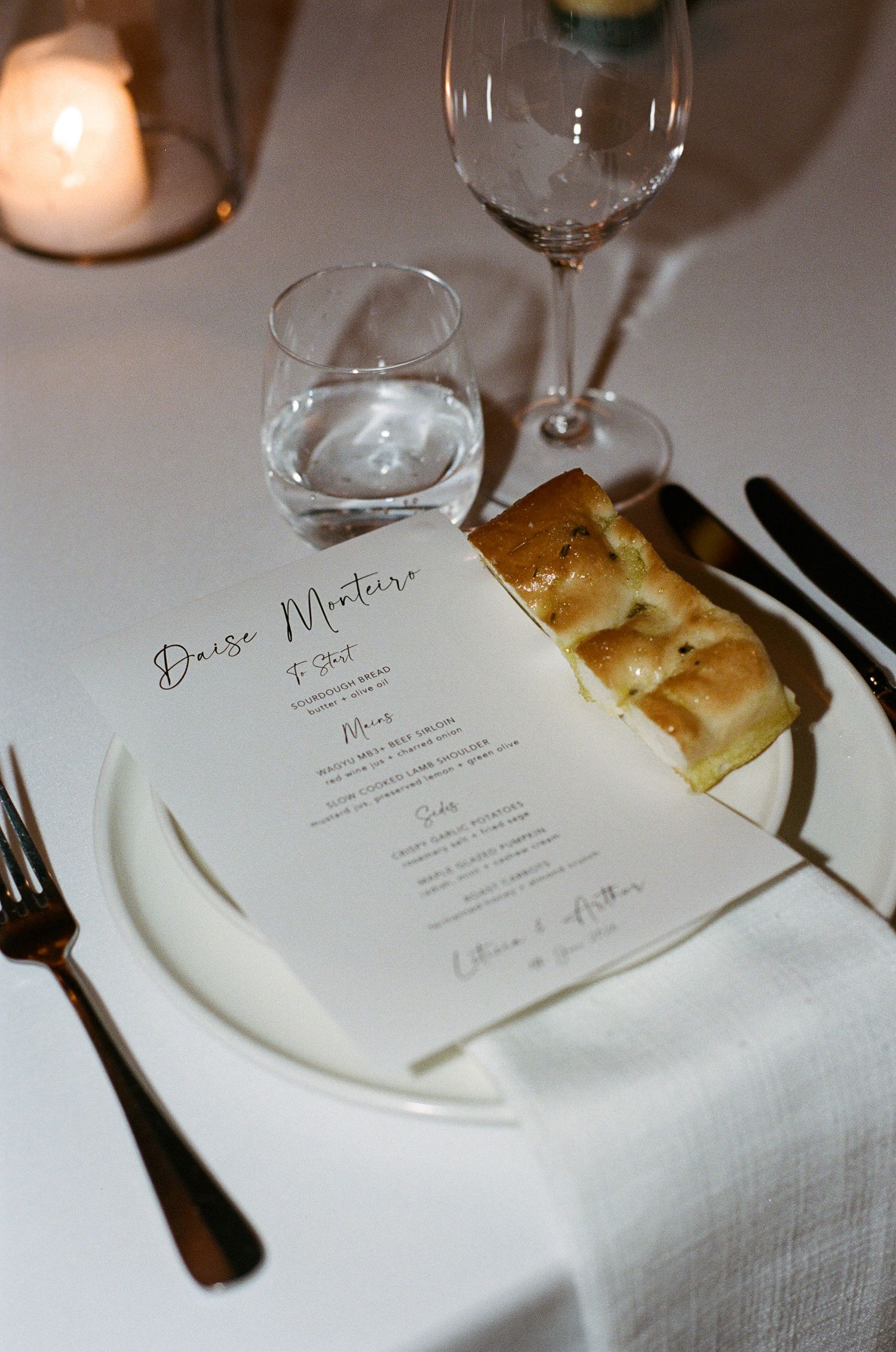 Elegant wedding table setting with a printed menu card, focaccia slice on a white plate, linen napkin, wine glass, water tumbler and candle.