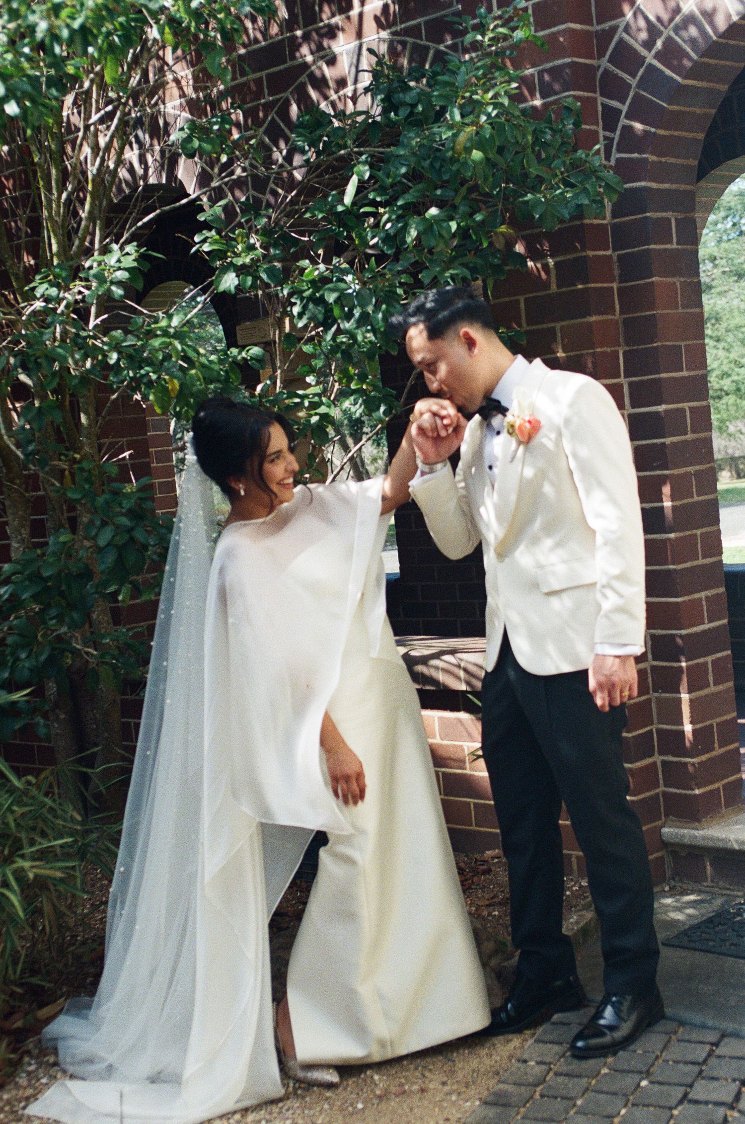 Groom in a white dinner jacket kisses the hand of a bride in a flowing white gown and long veil beside a brick archway.
