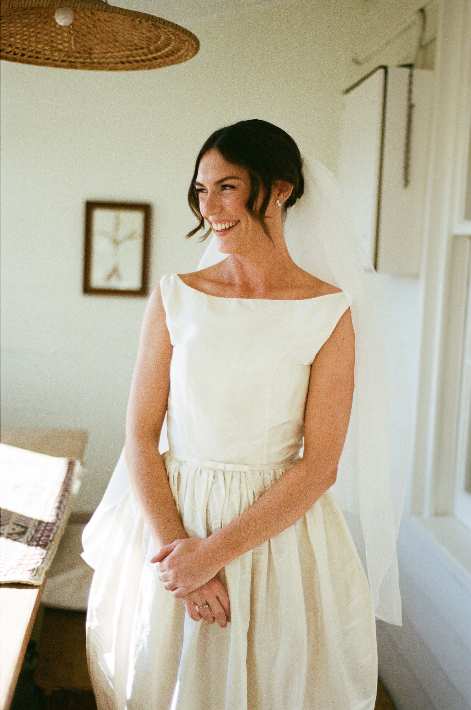 Smiling bride in a simple ivory sleeveless wedding dress and short veil stands with clasped hands in a bright, rustic room.