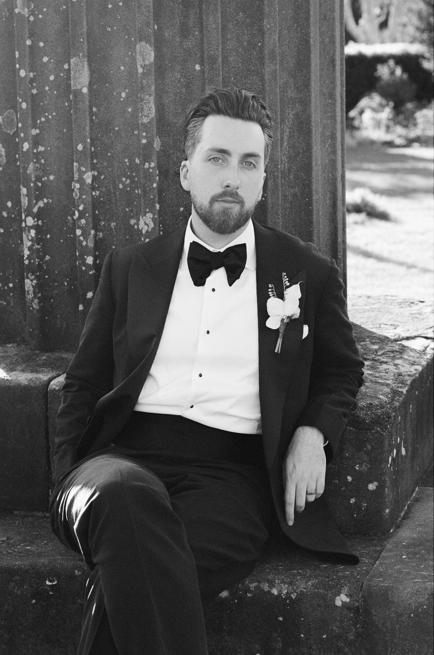 Black and white portrait of a bearded groom in a tuxedo and bow tie with a white floral boutonniere, seated on stone steps.