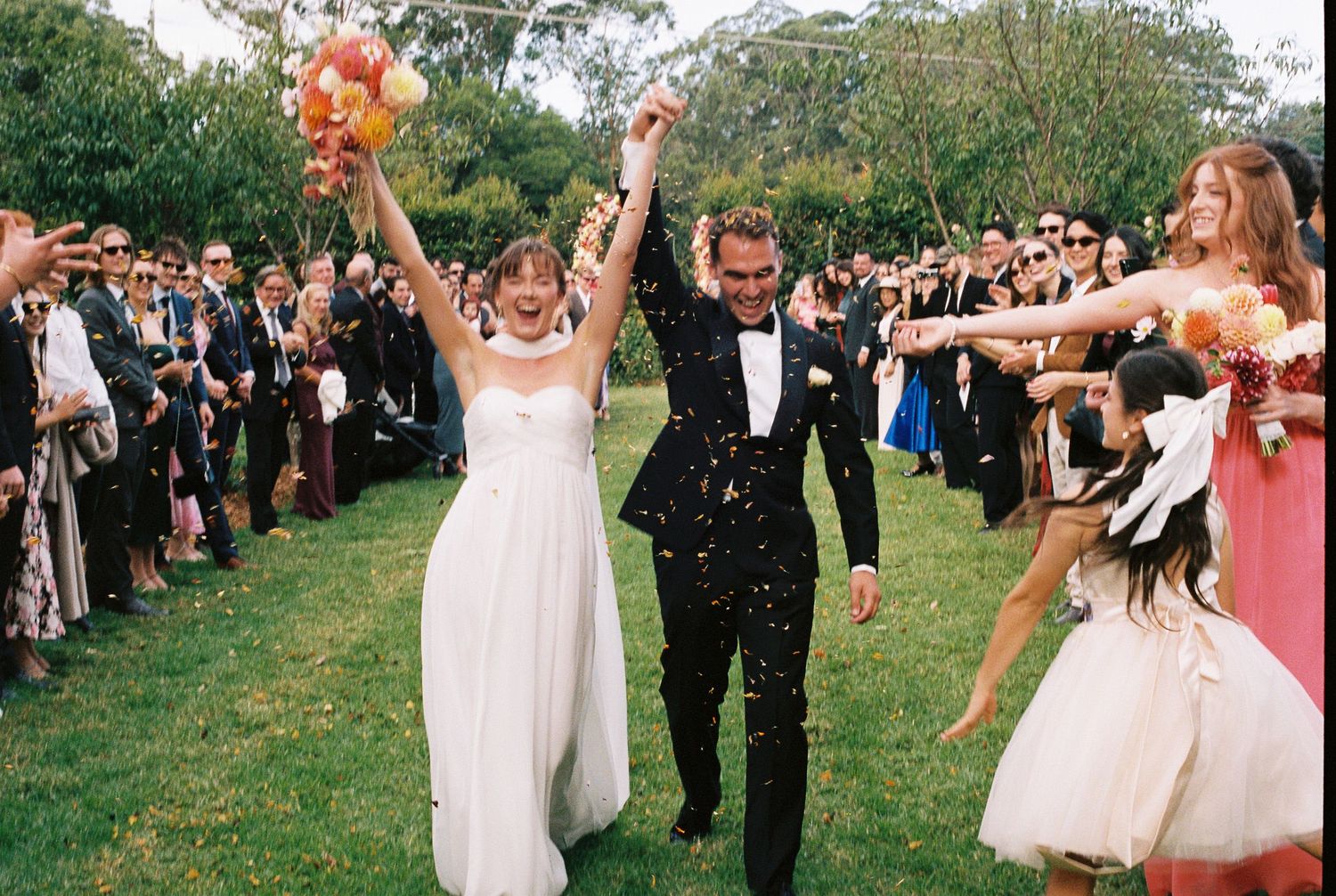 Joyful bride in a white gown and groom in a black suit walk hand in hand through cheering guests at an outdoor garden wedding.