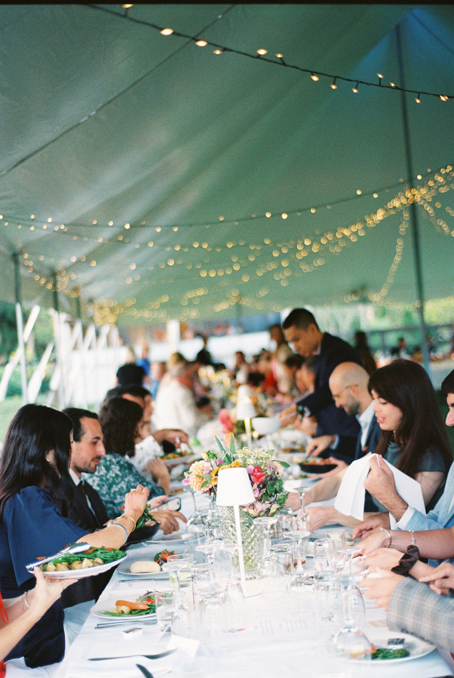 Guests enjoy a long communal table with fresh salads and flowers under a tent strung with warm twinkling lights at an outdoor reception.