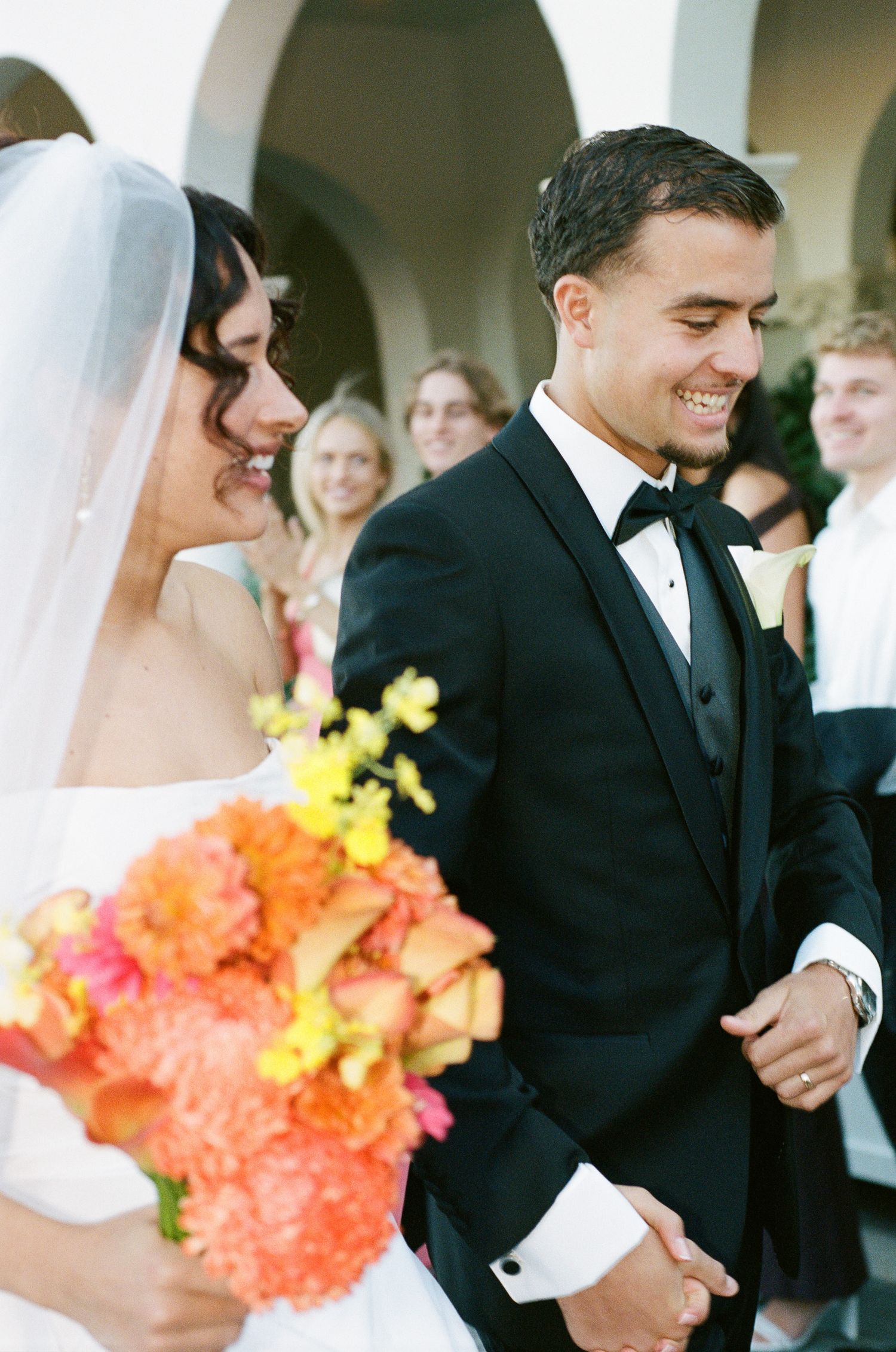 Smiling groom in a black tuxedo and bow tie walks beside a veiled bride holding an orange and pink bouquet at an outdoor wedding.