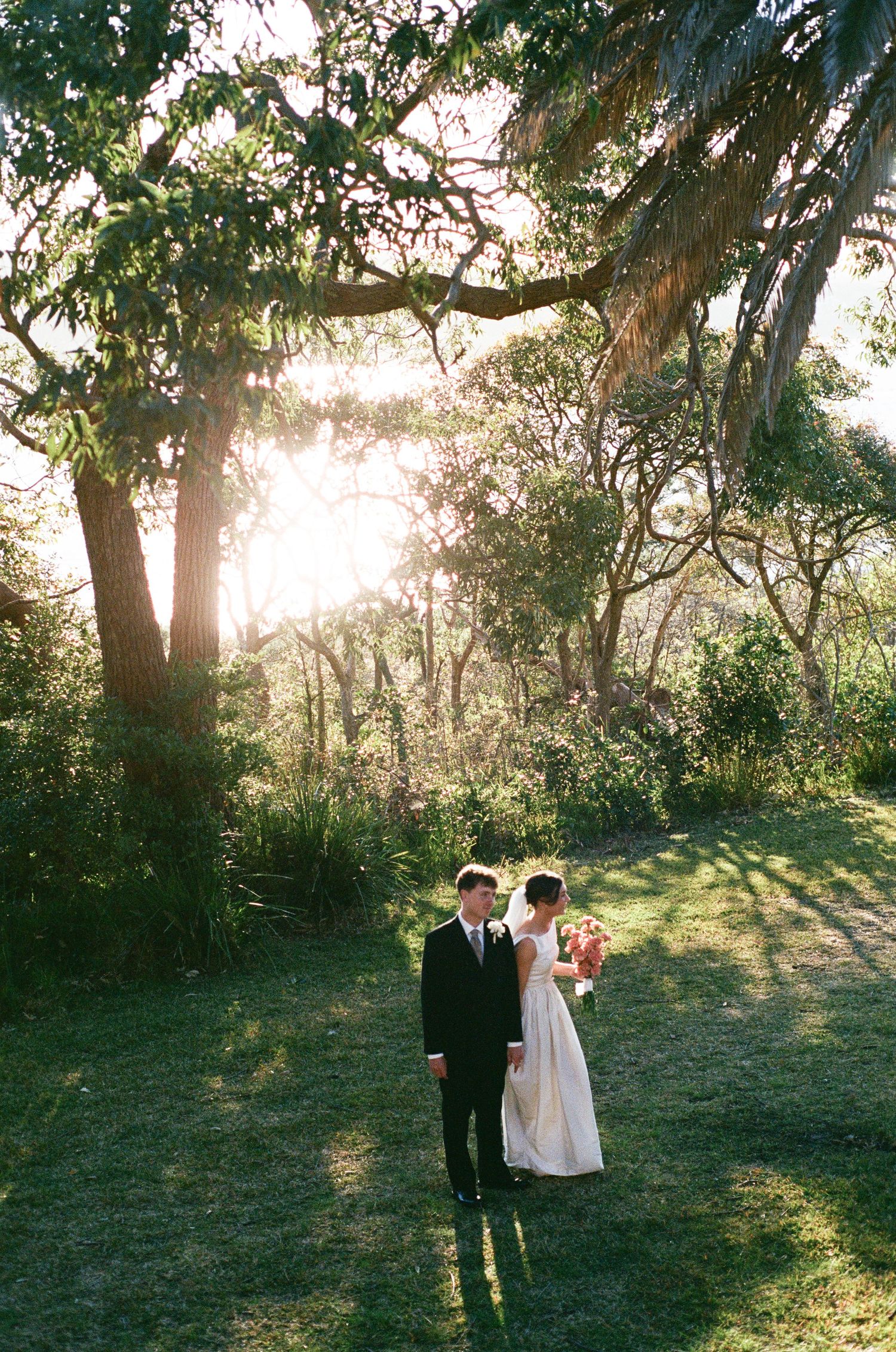 Bride and groom share a romantic moment in a sunlit garden surrounded by lush green trees.