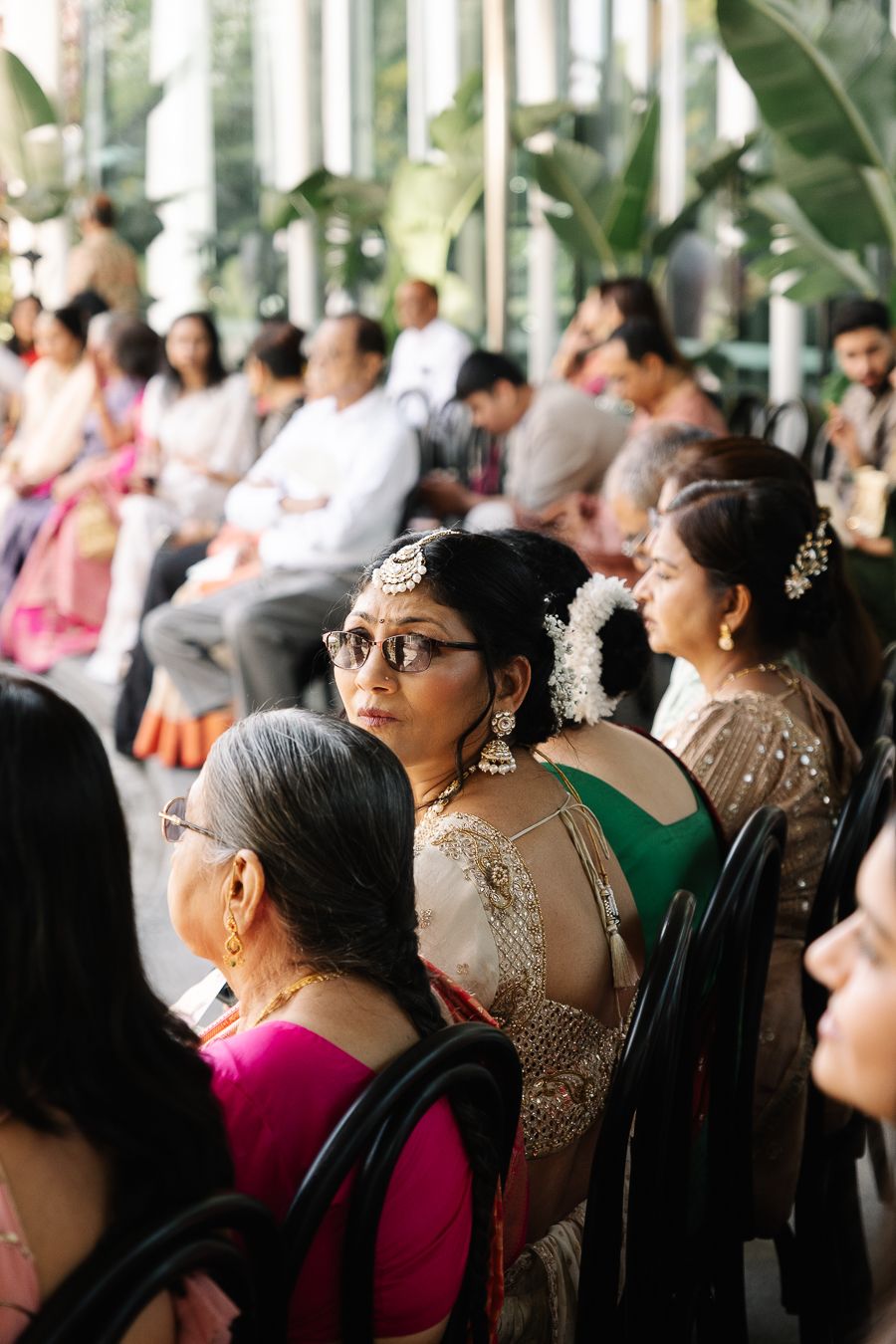 A crowd of people in formal attire attend an event, with decorative lighting and greenery visible in the background.