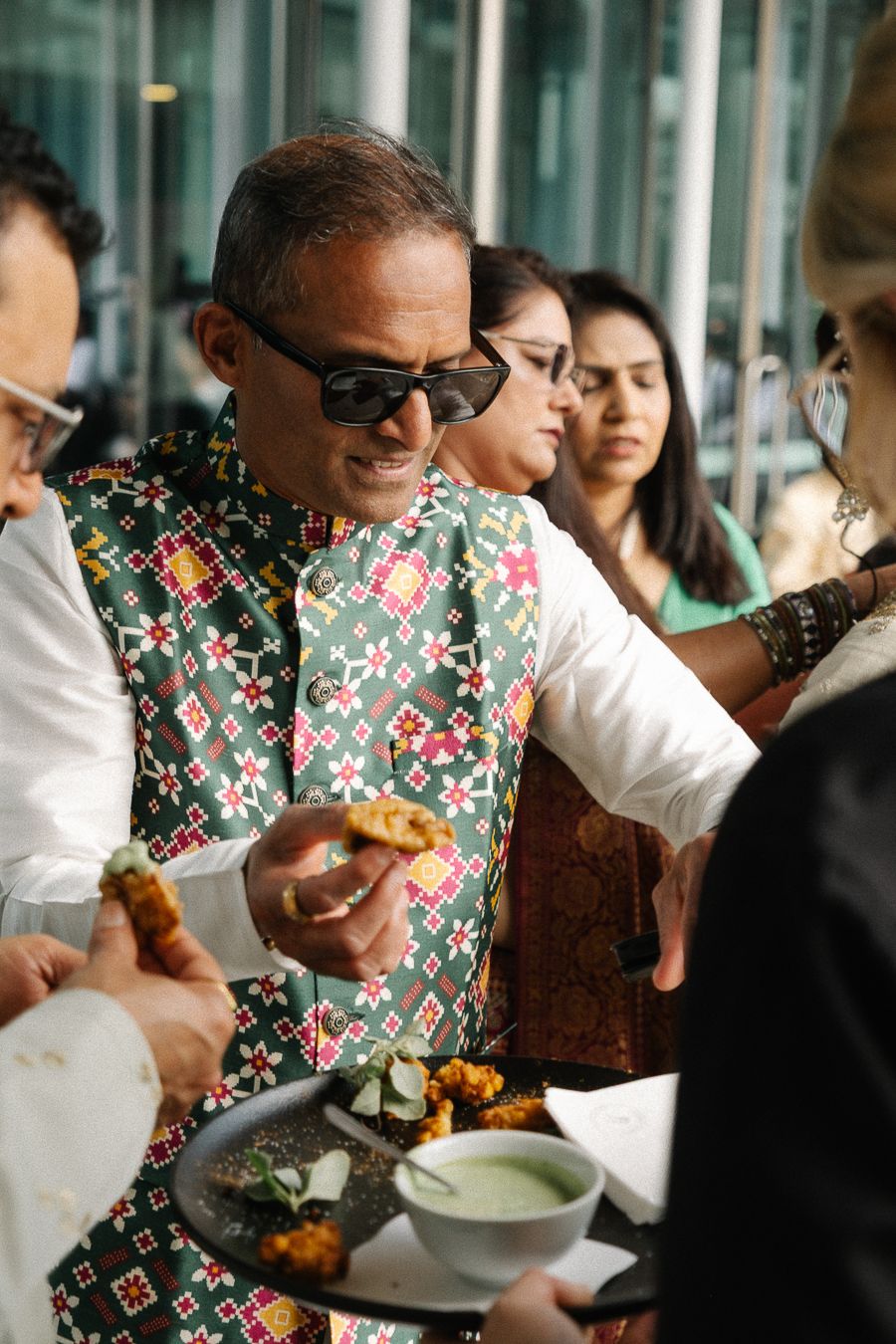 People gather at a social event wearing festive attire while eating and drinking together in a bright indoor space.
