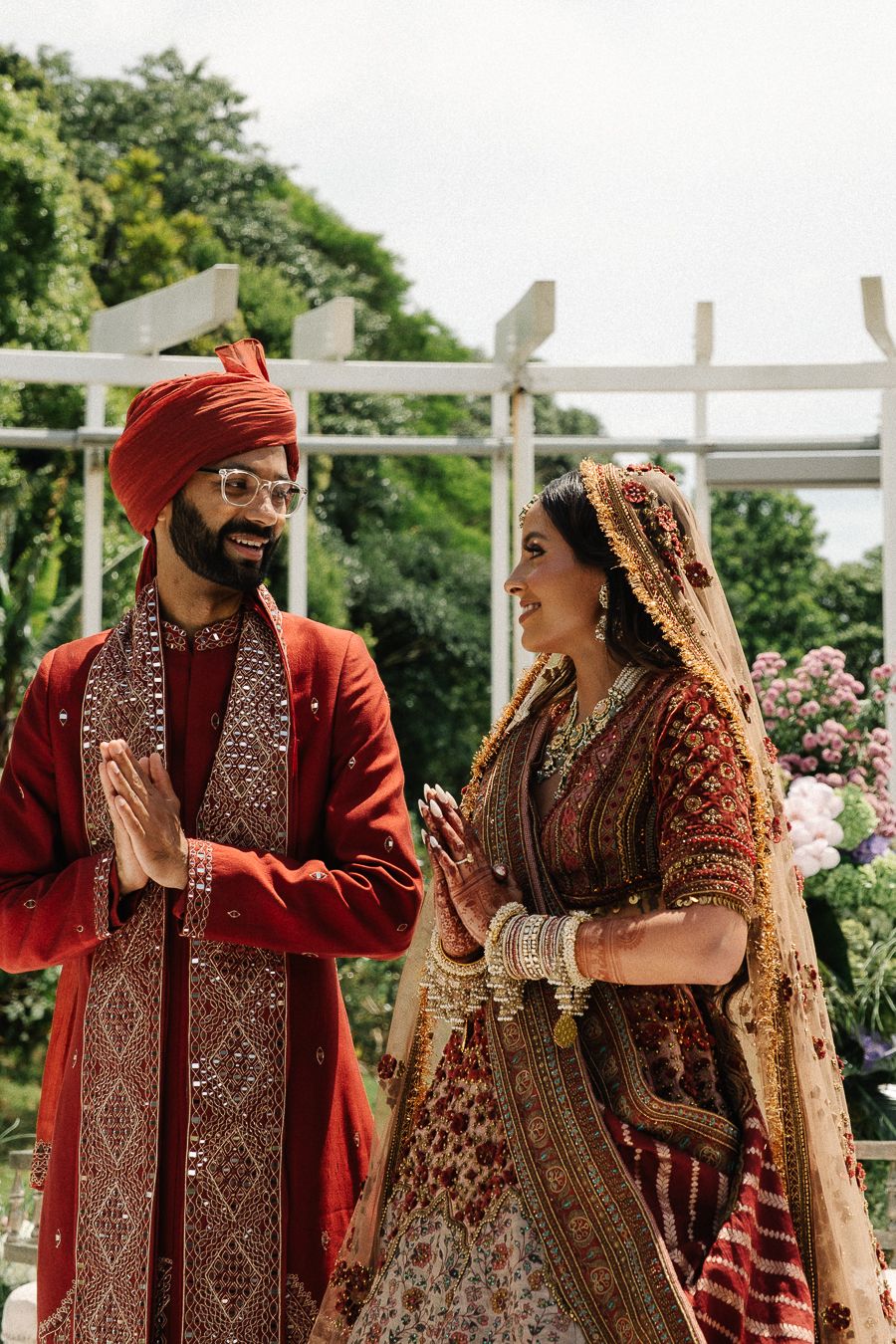A couple in traditional South Asian wedding attire share joyful moments in a garden setting with white lattice backdrop.