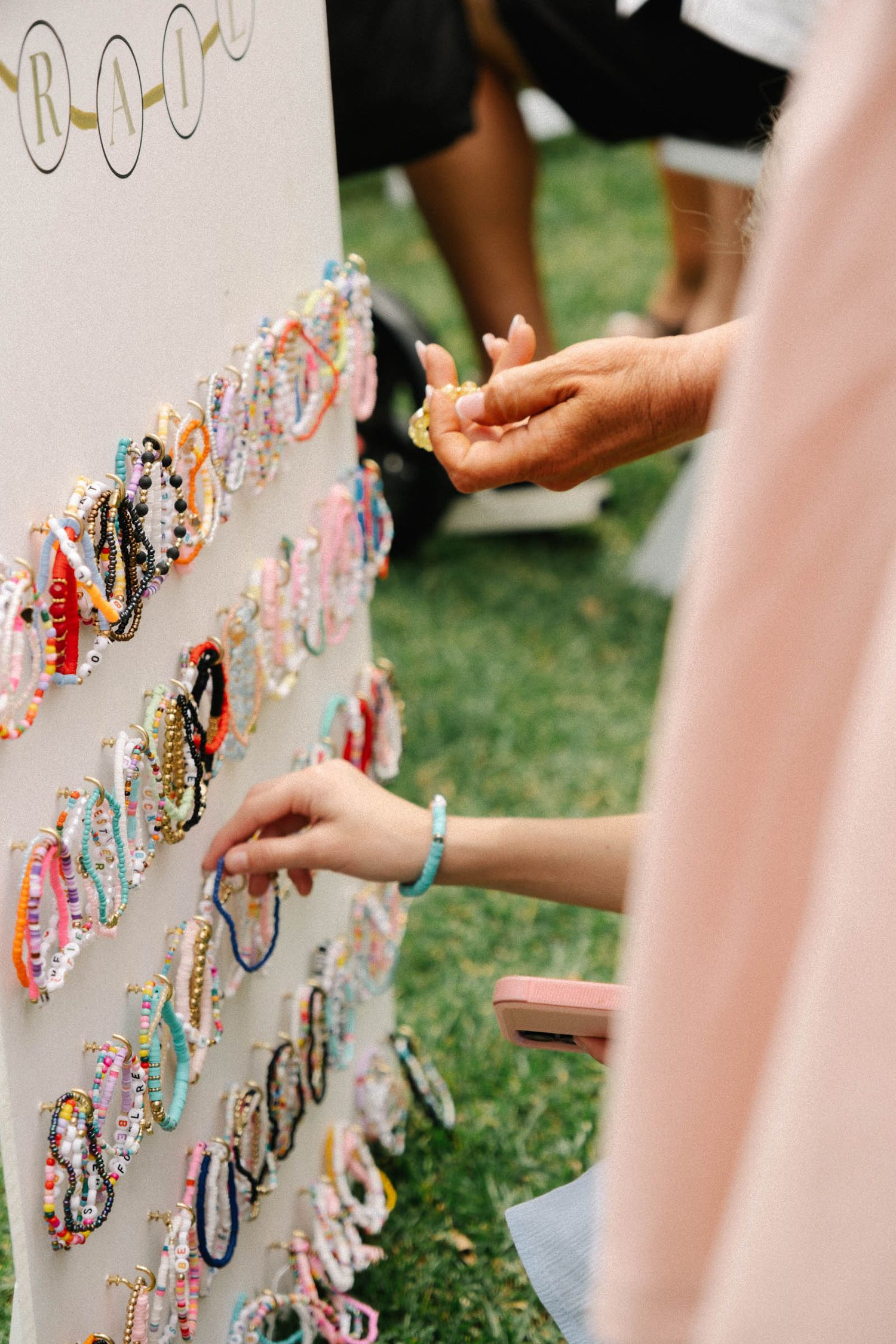 Colorful bracelets and jewelry displayed on a white stand at an outdoor event with hands reaching to browse items.