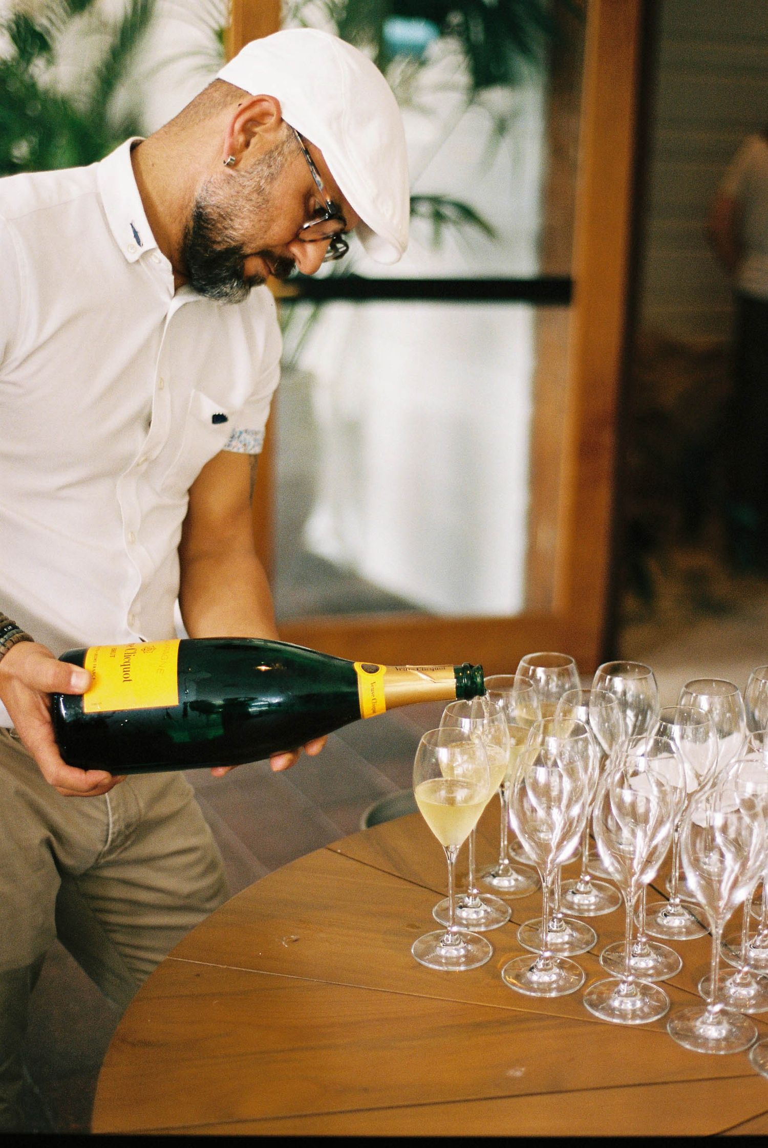A bartender in white shirt pours champagne into multiple wine glasses lined up on a wooden table.