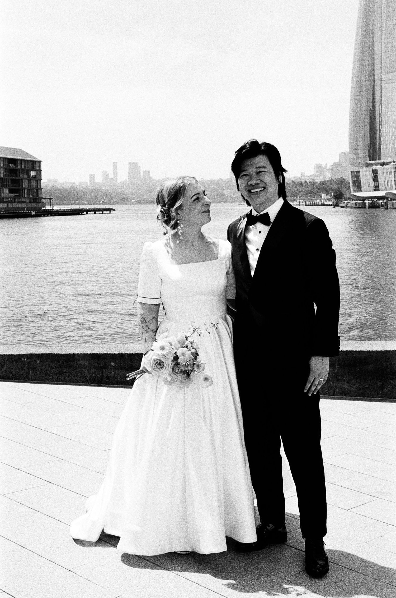 A black and white wedding photo series shows a couple posing together along a waterfront with a cityscape in the background.