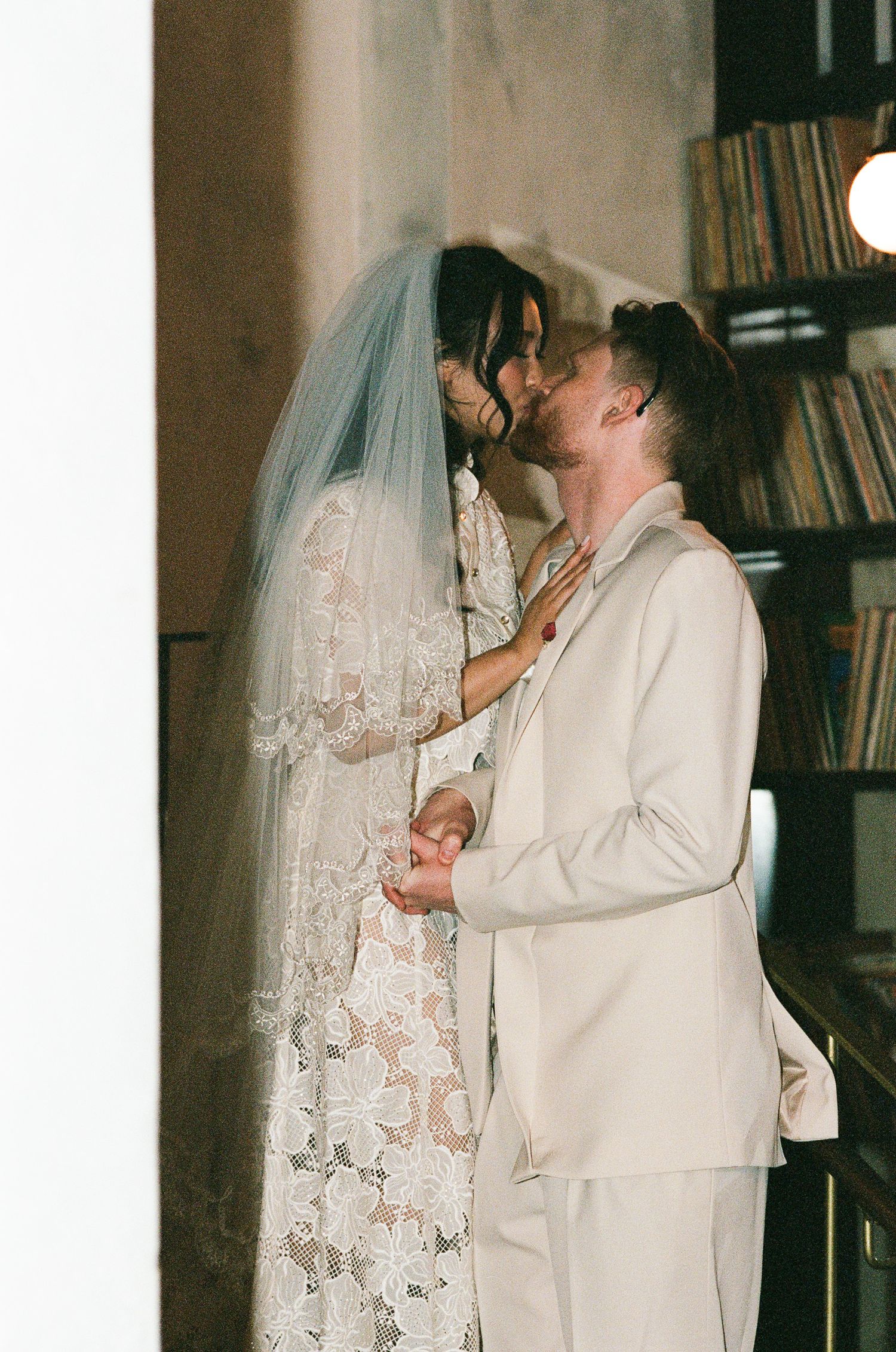 Bride in a lace gown and long veil kisses her groom in a cream suit beside a bookshelf in a warm, intimate wedding portrait.