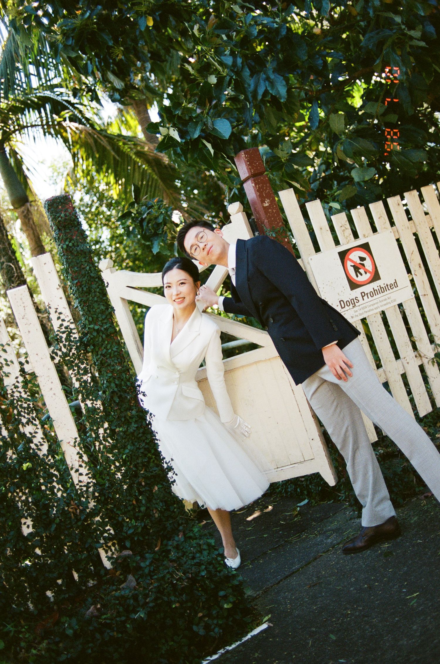 Bride in a white dress and groom in a navy blazer lean on a white picket gate surrounded by greenery near a Dogs Prohibited sign.