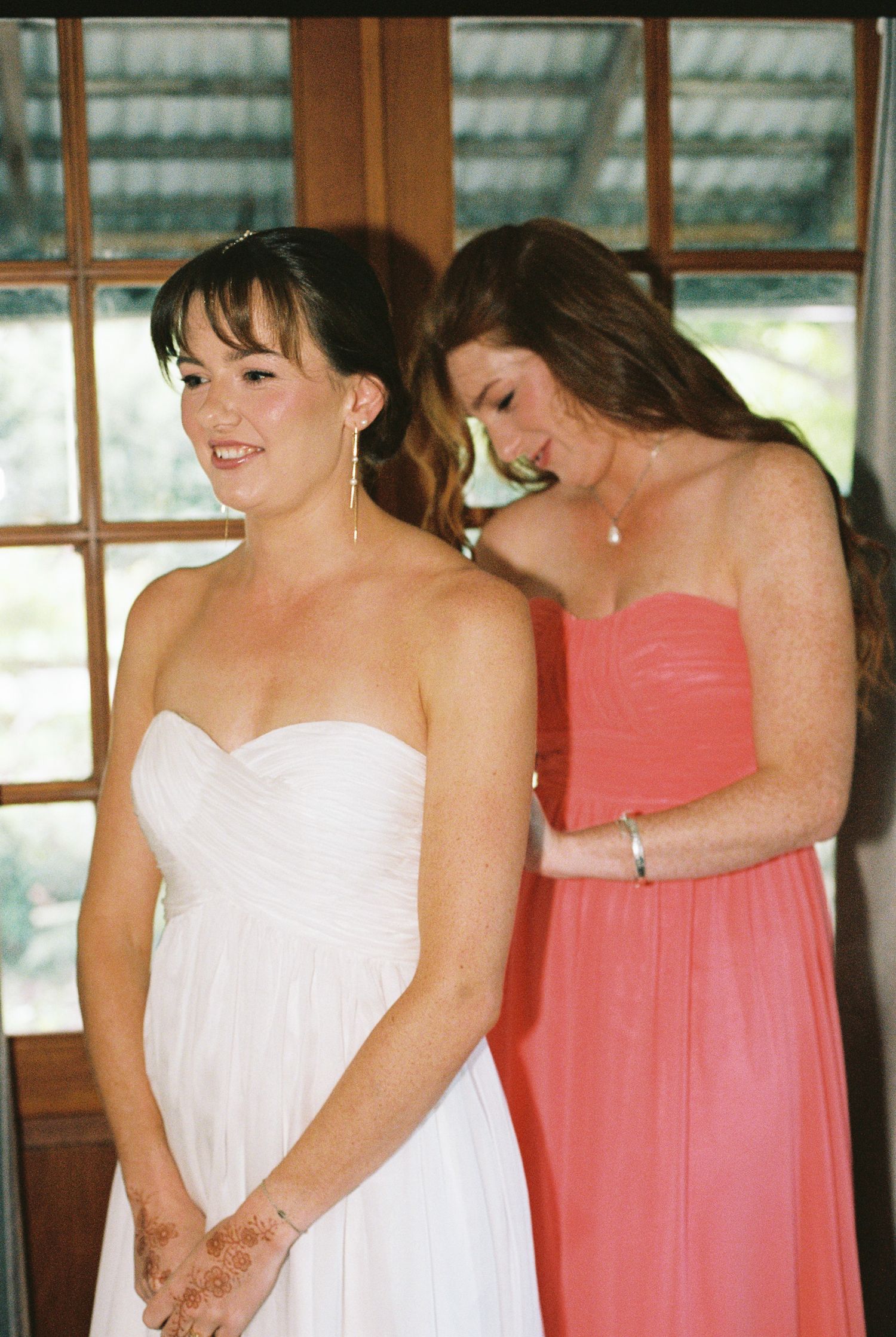 A bride in a white strapless gown with henna-decorated hands is helped into her dress by a bridesmaid in a coral pink dress.