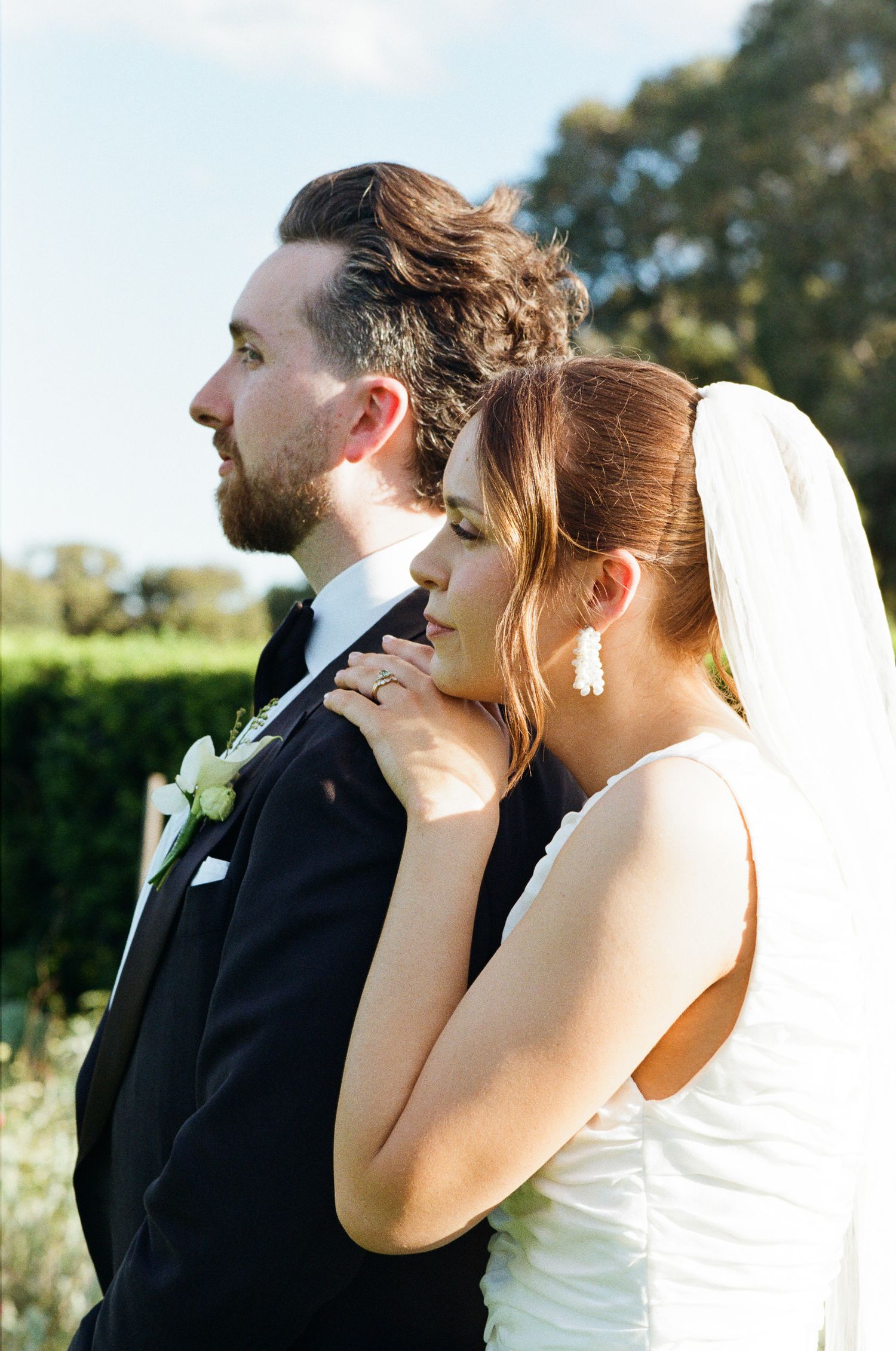 Bride in a white gown and veil rests her hand on the groom's shoulder as the couple gazes into the distance in a sunny garden.