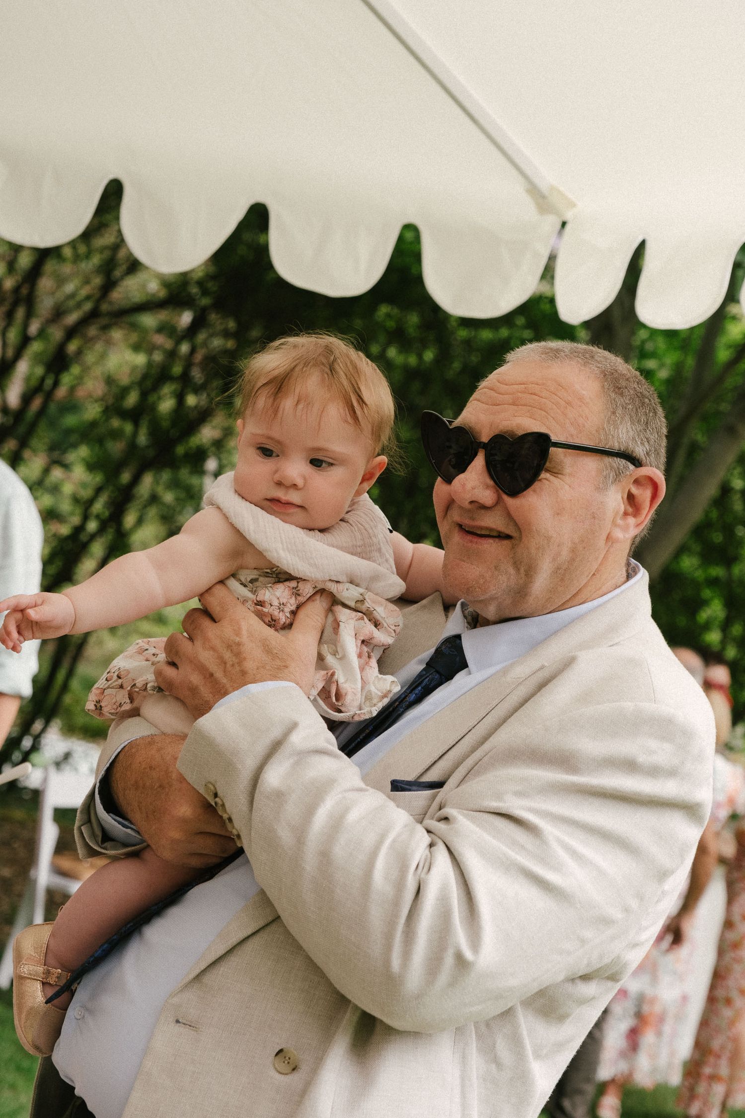 A baby is held and entertained under a white canopy at an outdoor gathering.