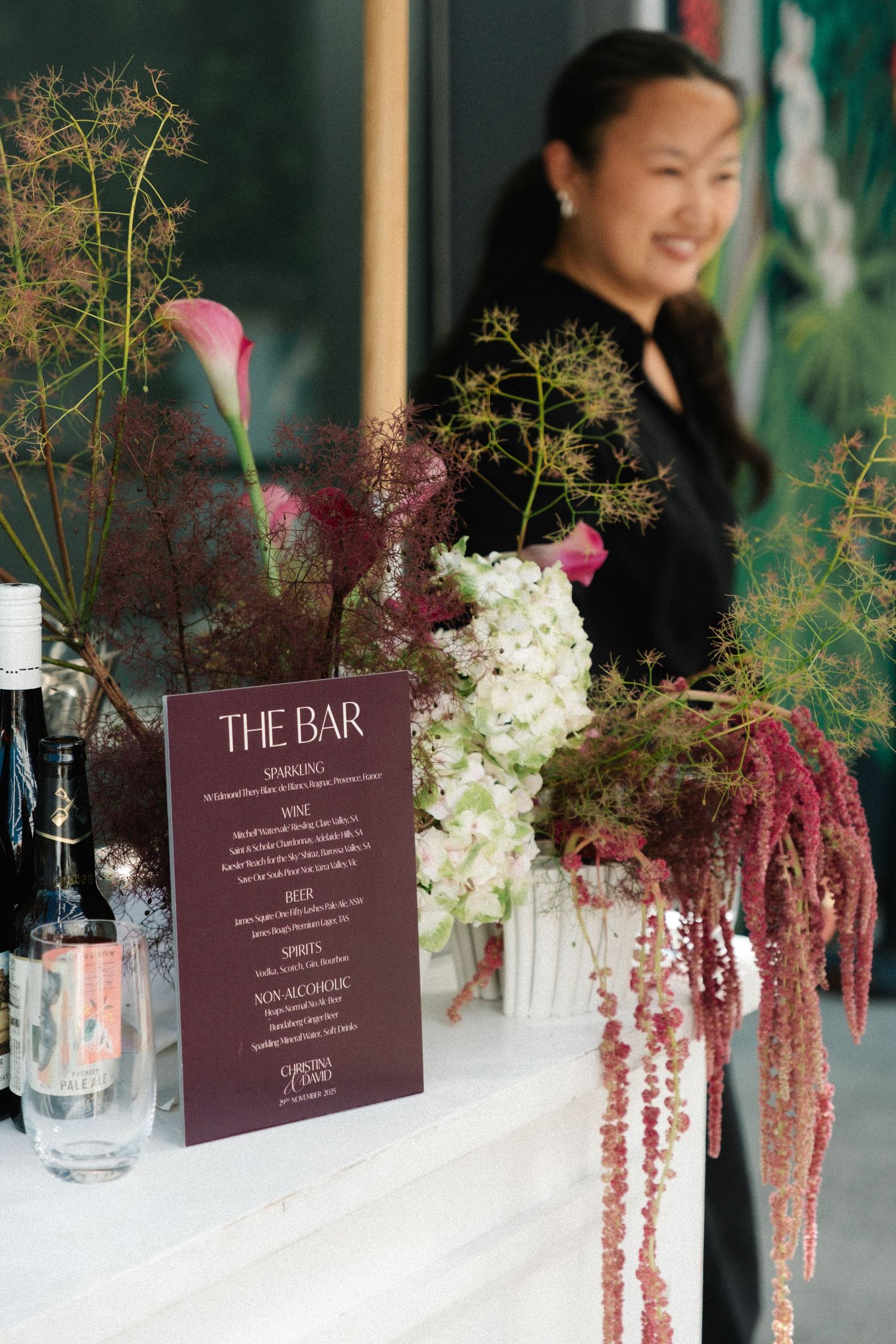 A bar menu stands on a white table with elegant floral arrangements featuring white hydrangeas and cascading burgundy amaranth.