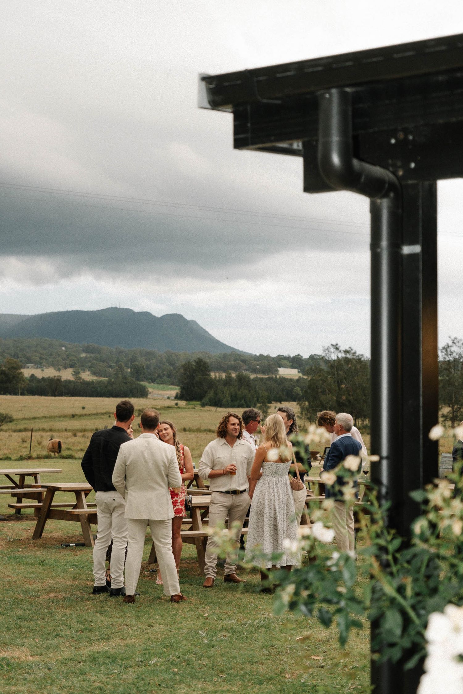 Outdoor wedding ceremony setup with white-clad guests gathered under covered pavilion overlooking mountain landscape.