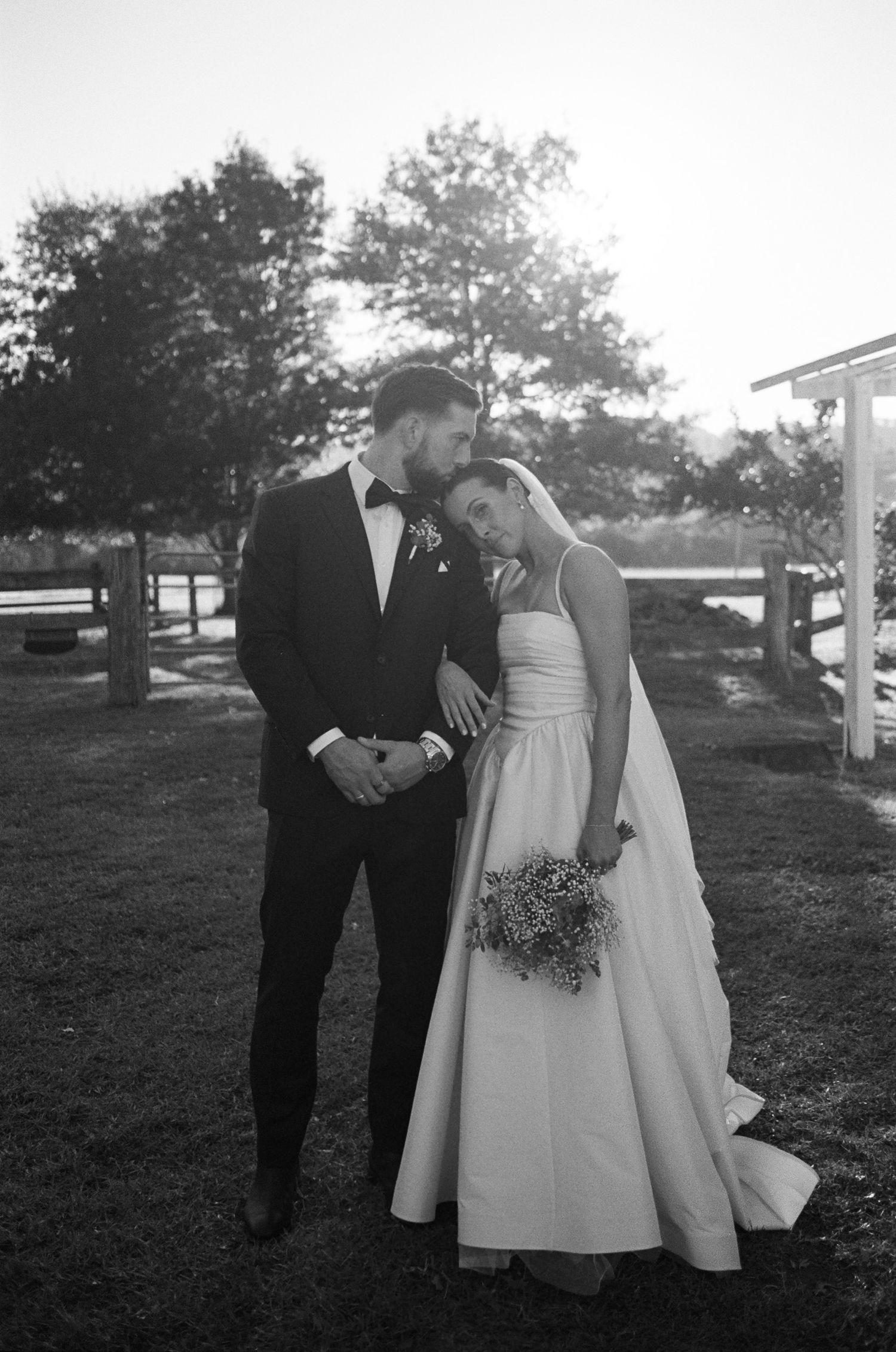 Black and white photo of a bride in a strapless gown resting her head on the groom's shoulder, holding a bouquet outdoors at golden hour.