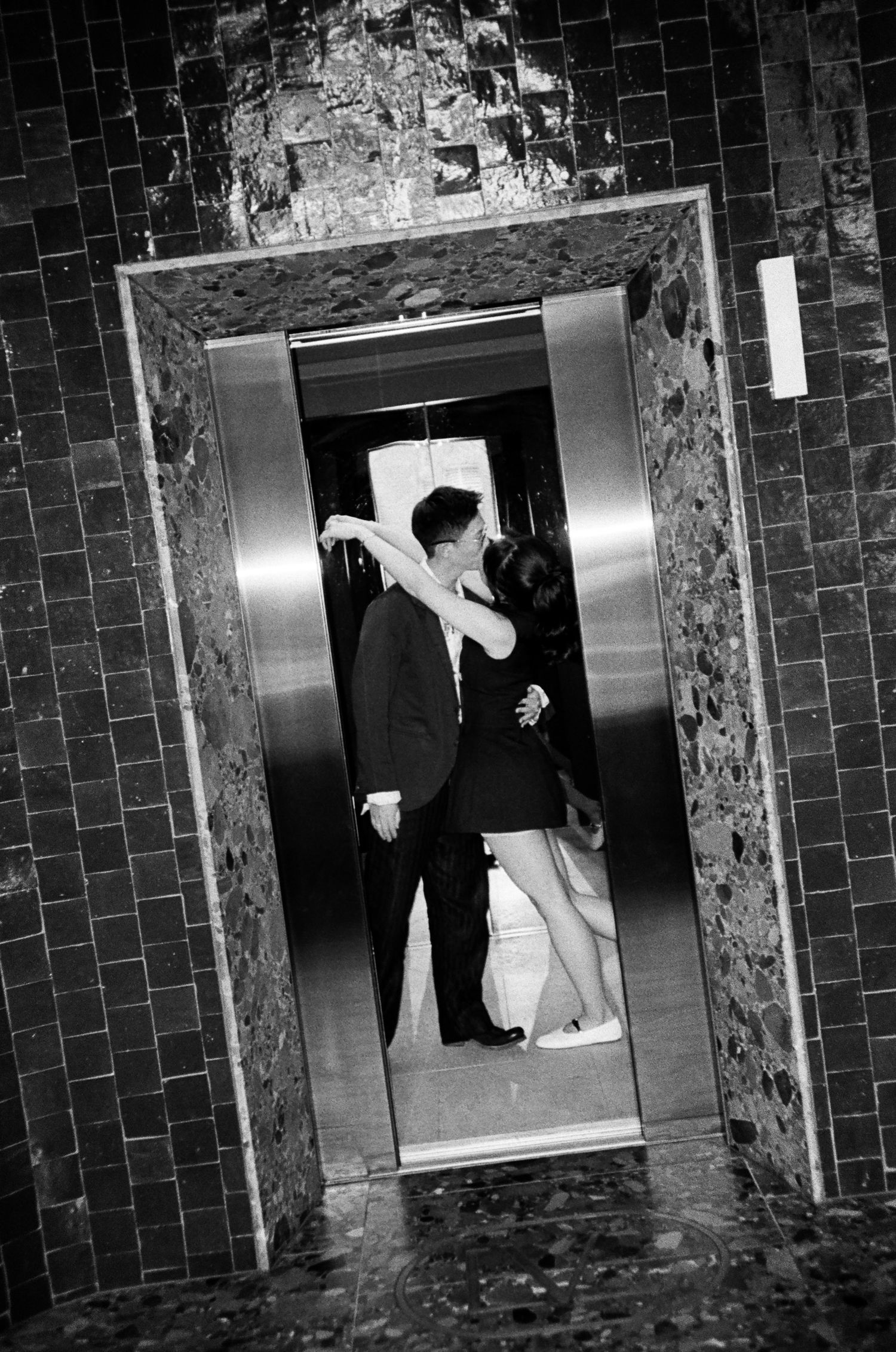 Black and white photo of a couple kissing inside an open elevator framed by dark tiled walls and a stone-patterned entrance.