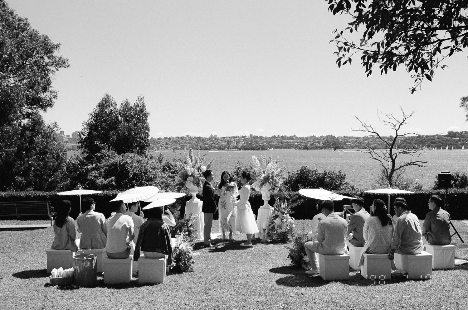 Black and white photo of an outdoor wedding ceremony on a grassy lawn with seated guests, white parasols, and a couple before rolling hills.