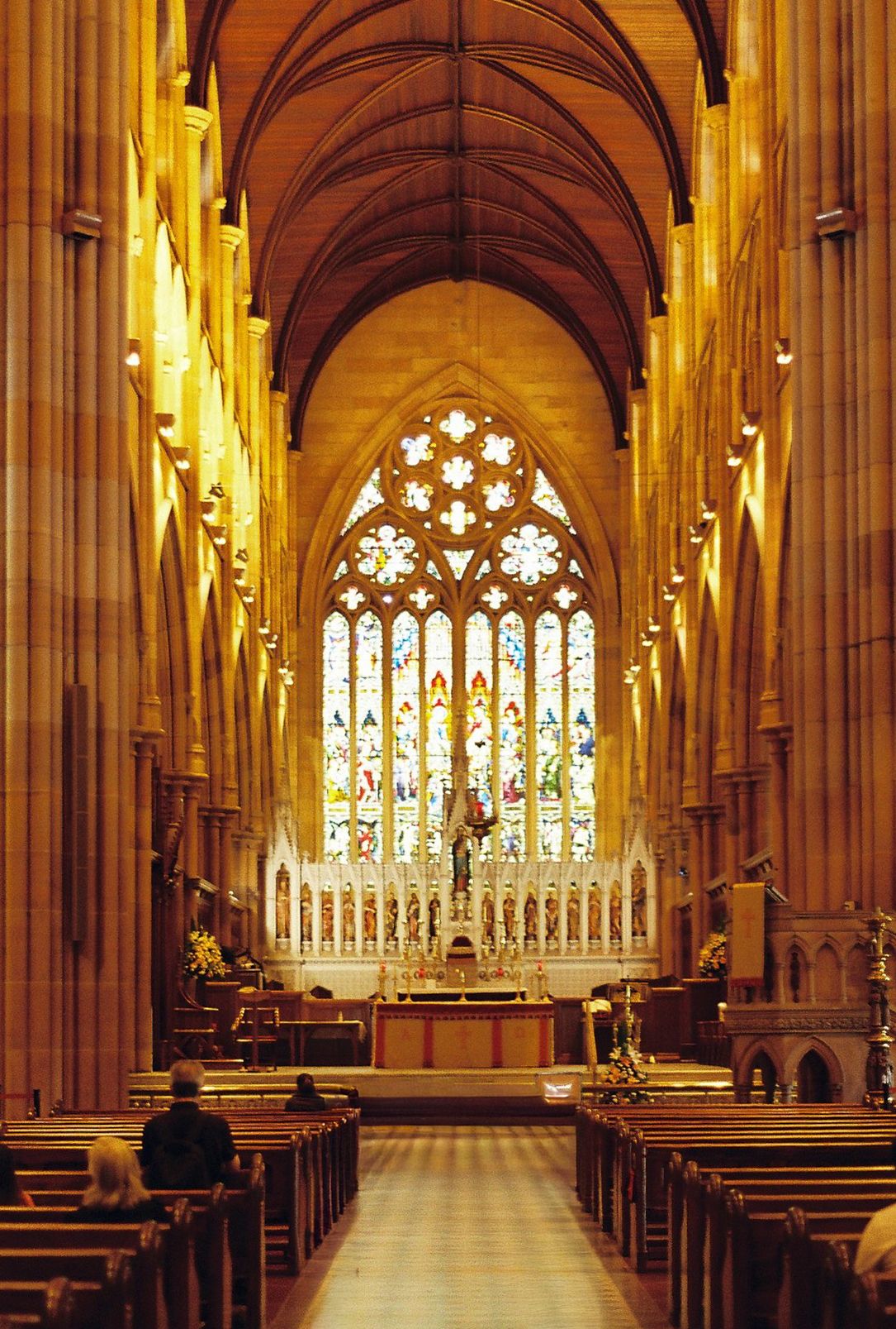 Interior view of a Gothic cathedral with vaulted ceiling, wooden pews, and large stained glass window at the altar.