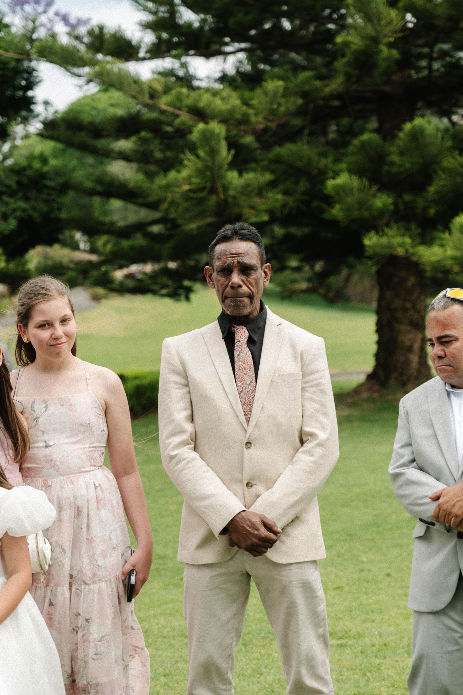 Wedding party members in cream colored suits and floral dress pose together on grassy lawn with pine trees in background.